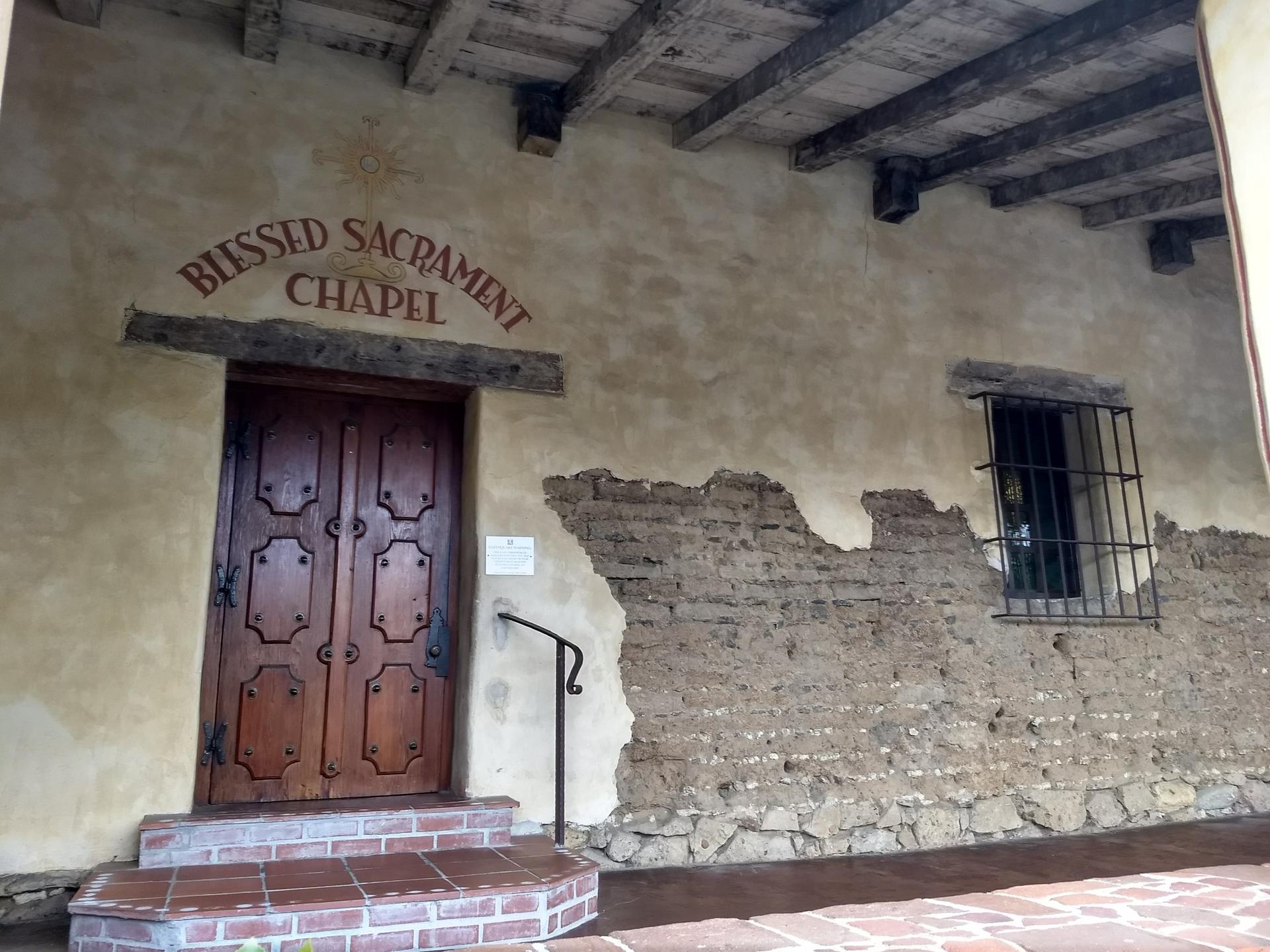 An old chapel entrance with a wooden door and exposed adobe wall.