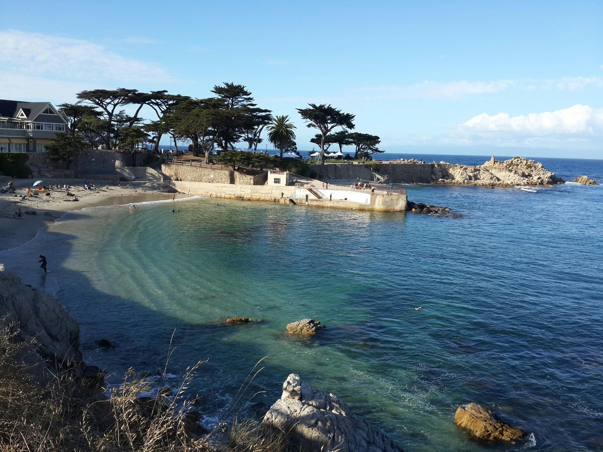 Sunny coastal cove with a sandy beach, clear water, and distant buildings.