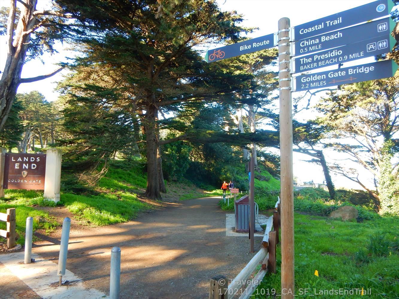 Dirt path through sunny woods with a directional sign and distant cyclist.