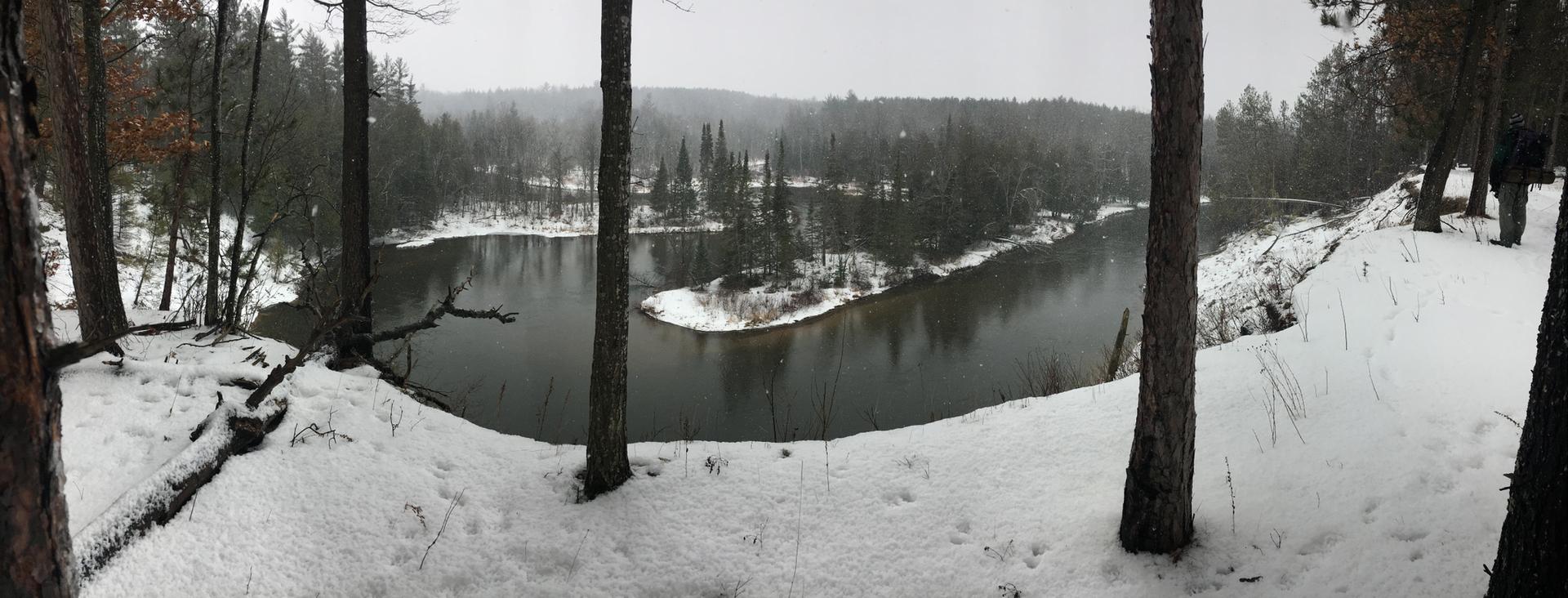 Panoramic view of a winding river through a snowy forest on an overcast day.