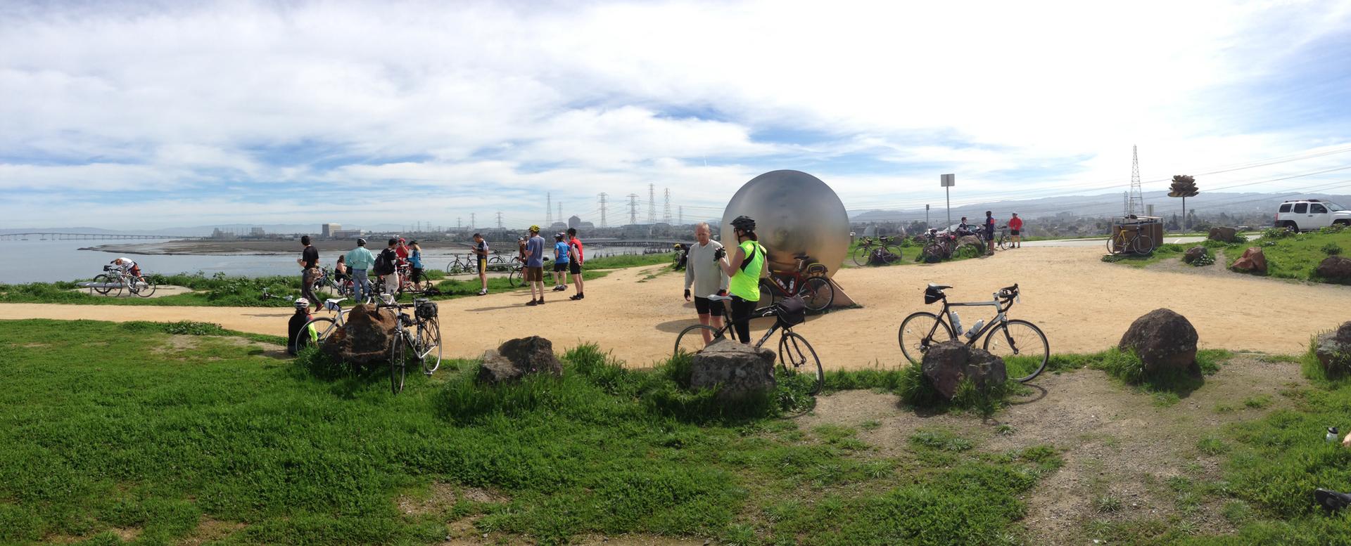 Group of people and bicycles gathered near a large sphere by the water.