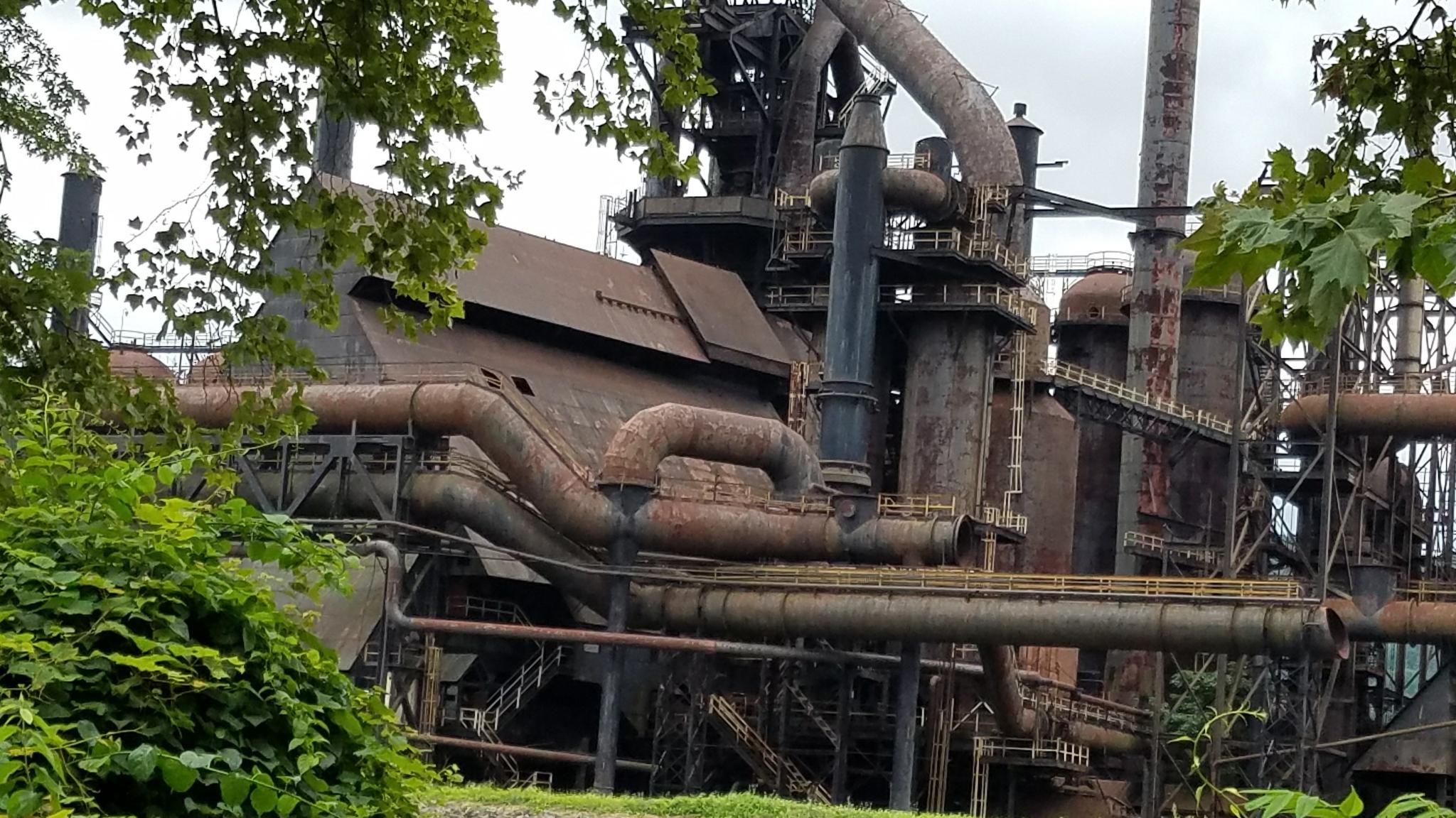 Rusting industrial complex with large pipes and structures, partially obscured by green foliage.