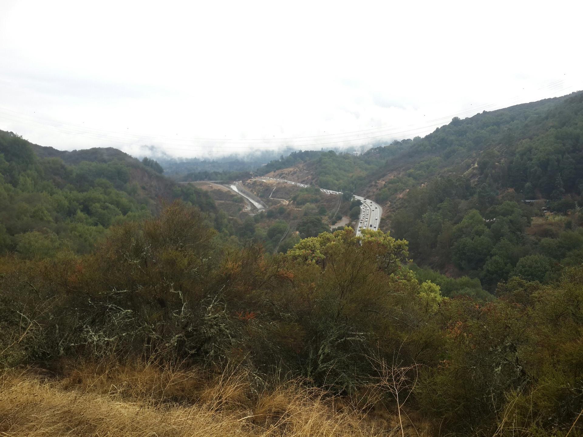 A winding road cuts through a forested valley under an overcast sky.