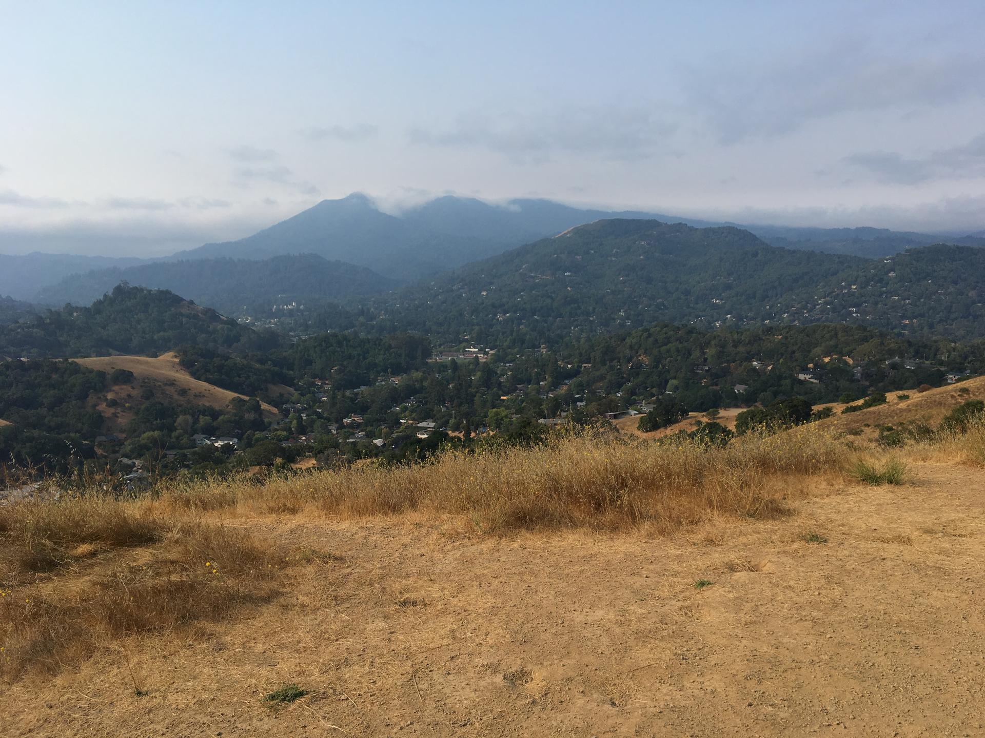 Expansive view of dry hills, green valleys, and distant mountains under a cloudy sky.