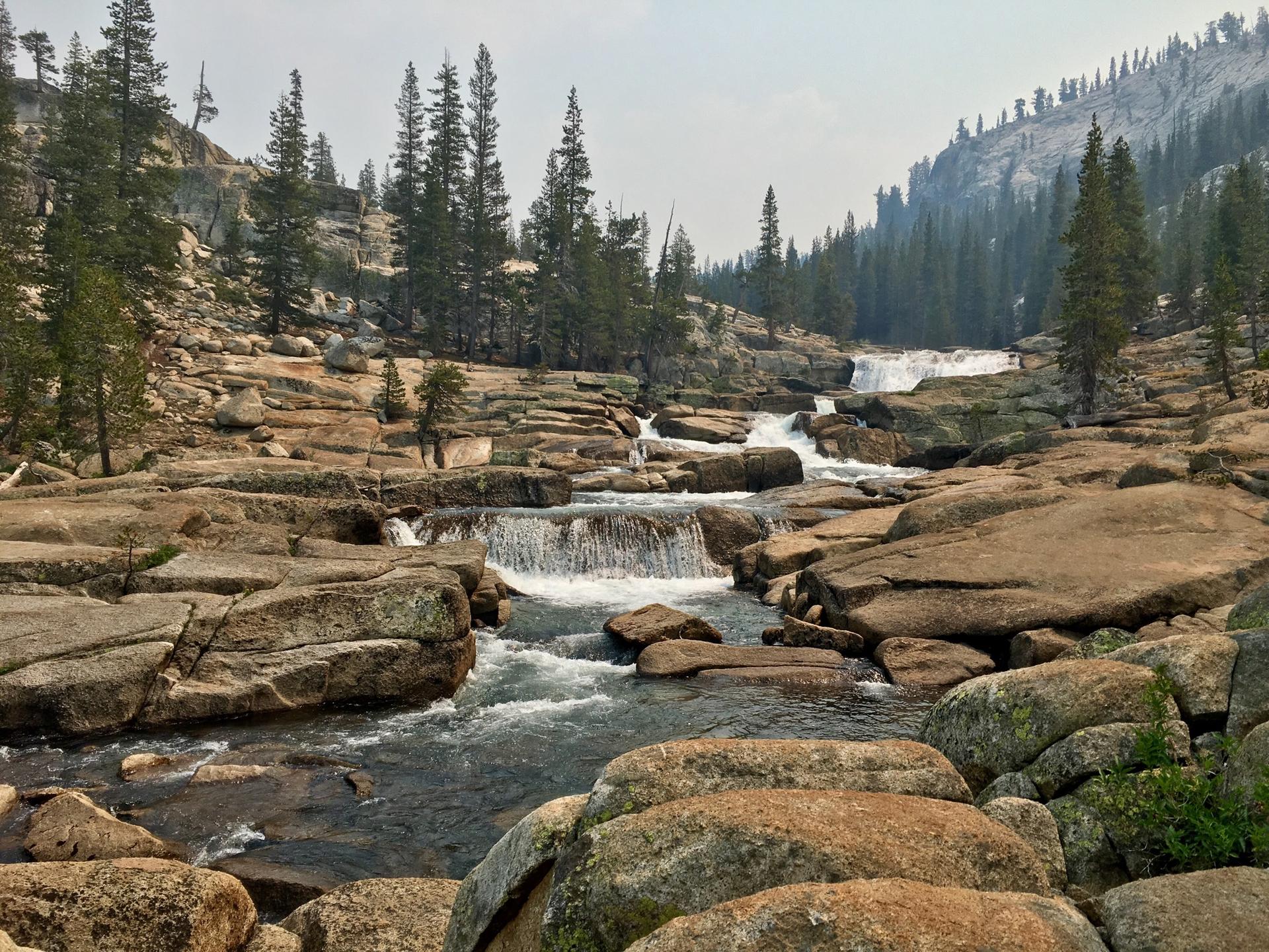 A river cascades over multiple rocky ledges through a pine forest on a hazy day.