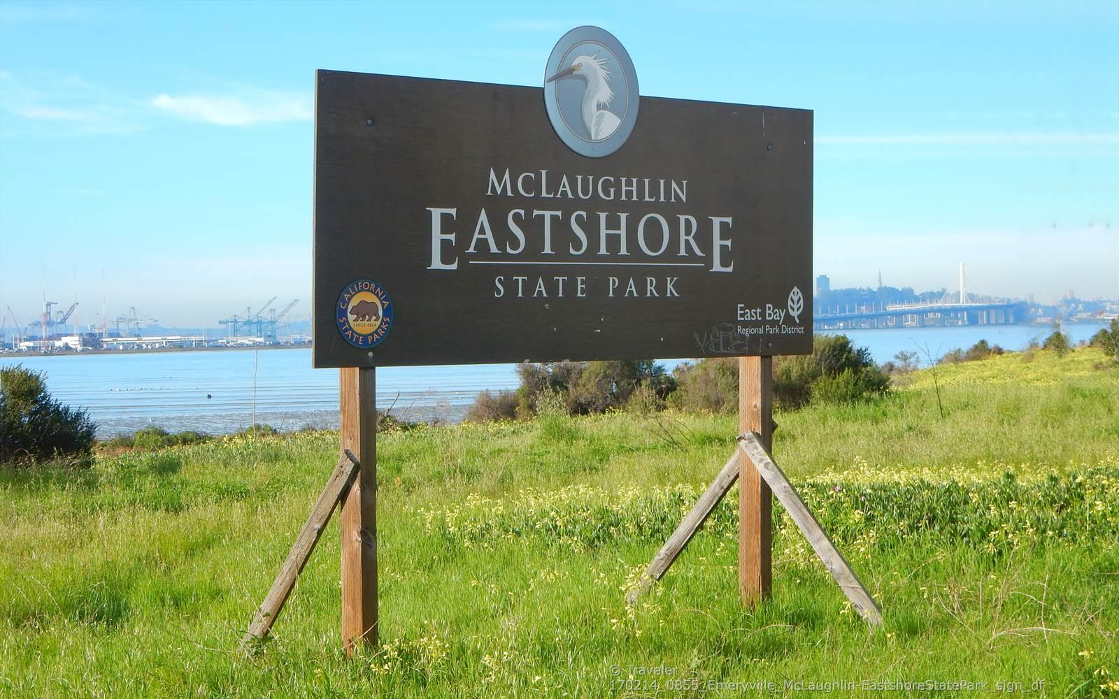 Park sign in a grassy field with a bay and distant city.