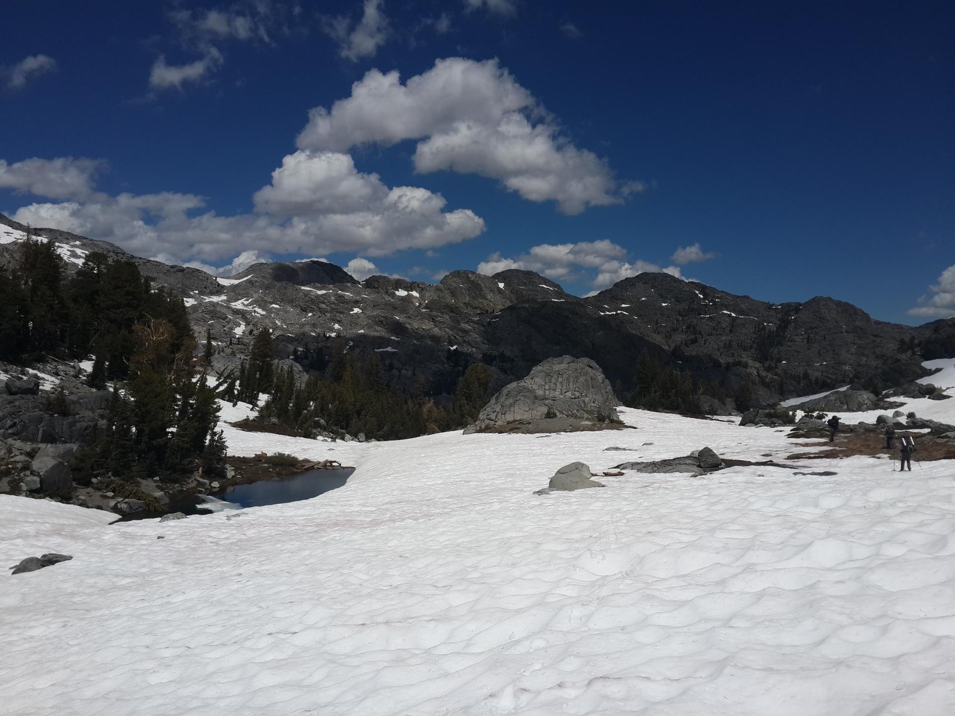 Bright, sunny mountain landscape with snow, rocks, and a distant person.