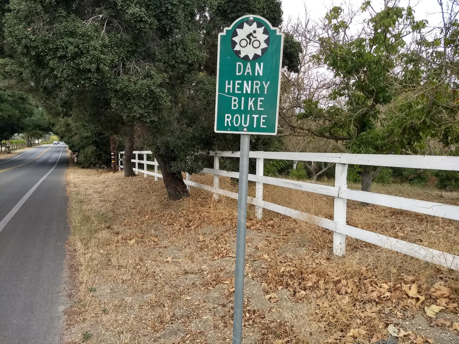 Green bike route sign next to a road and white fence.