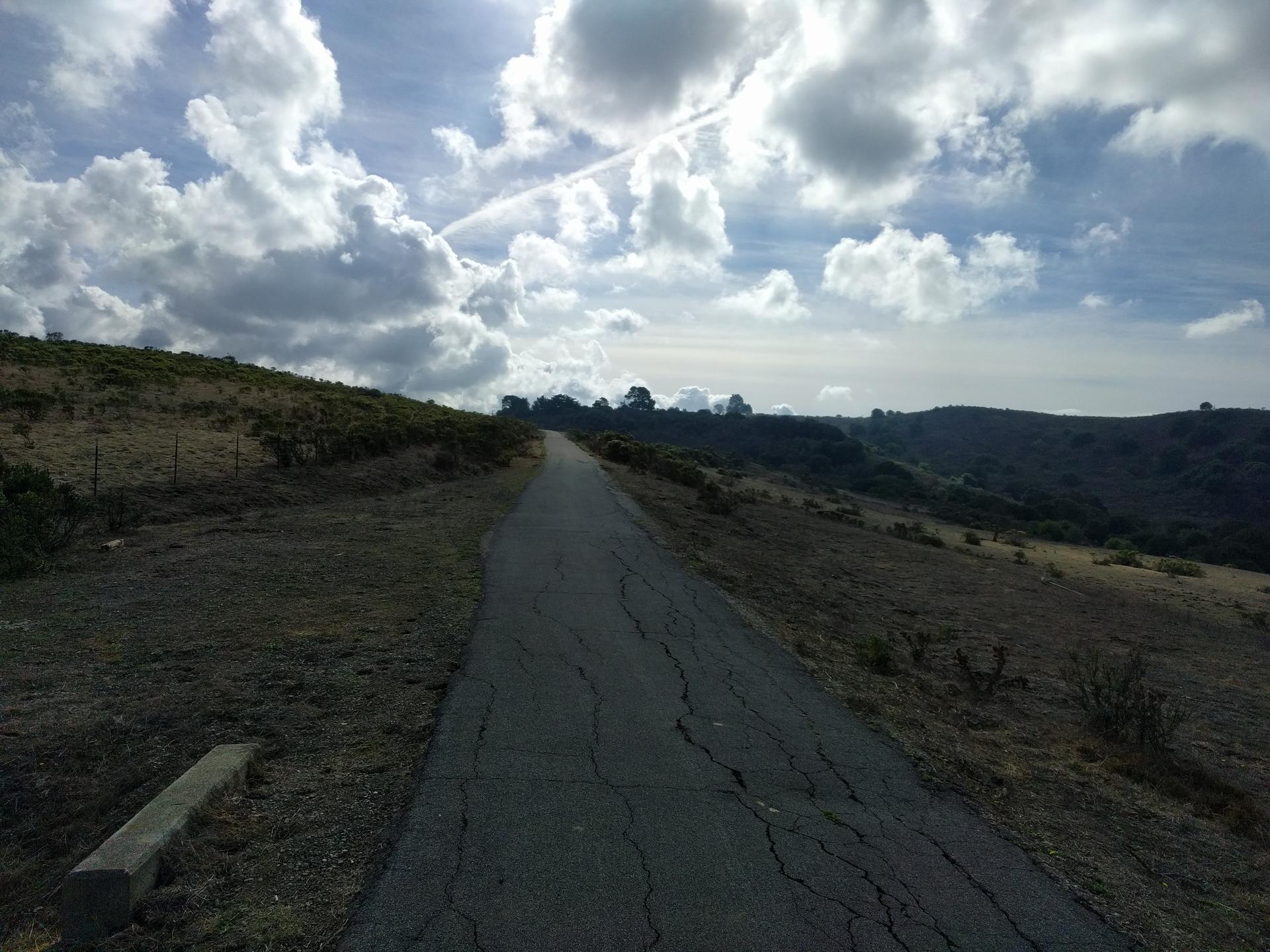 Paved path winding uphill through grassy hills under a cloudy sky.