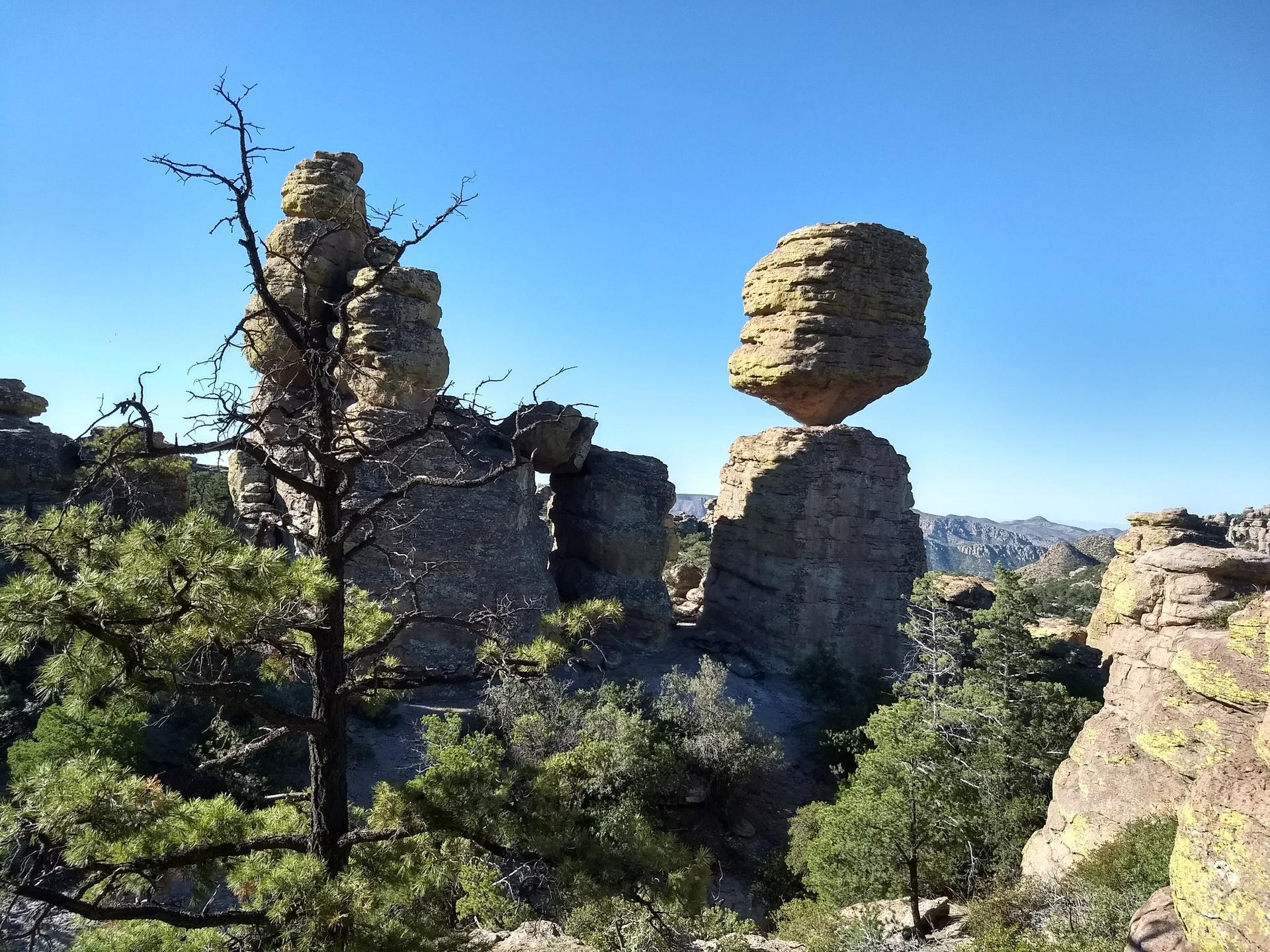 A large balanced rock formation and trees under a clear blue sky.