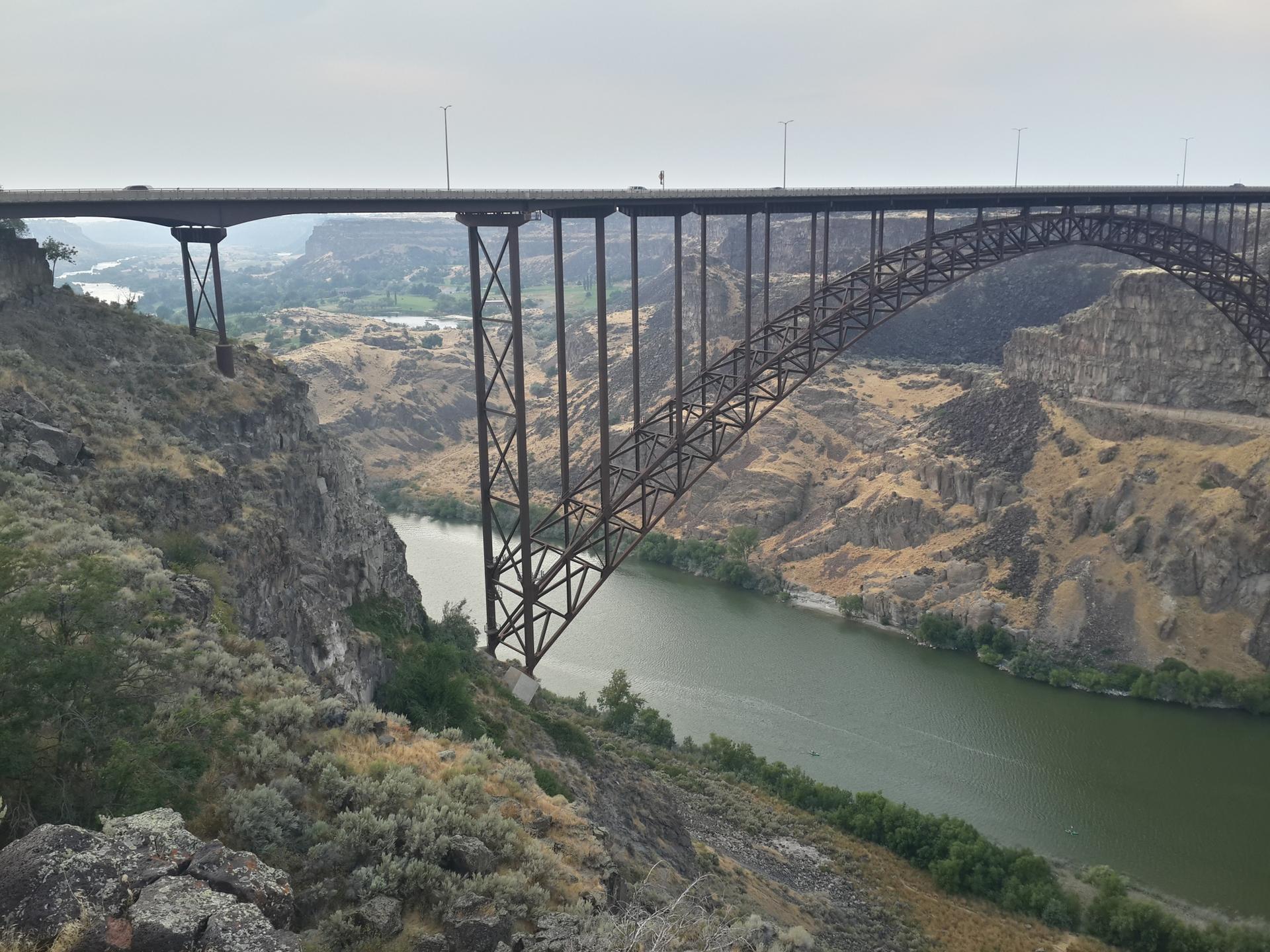 Large steel arch bridge spans a deep canyon river under an overcast sky.