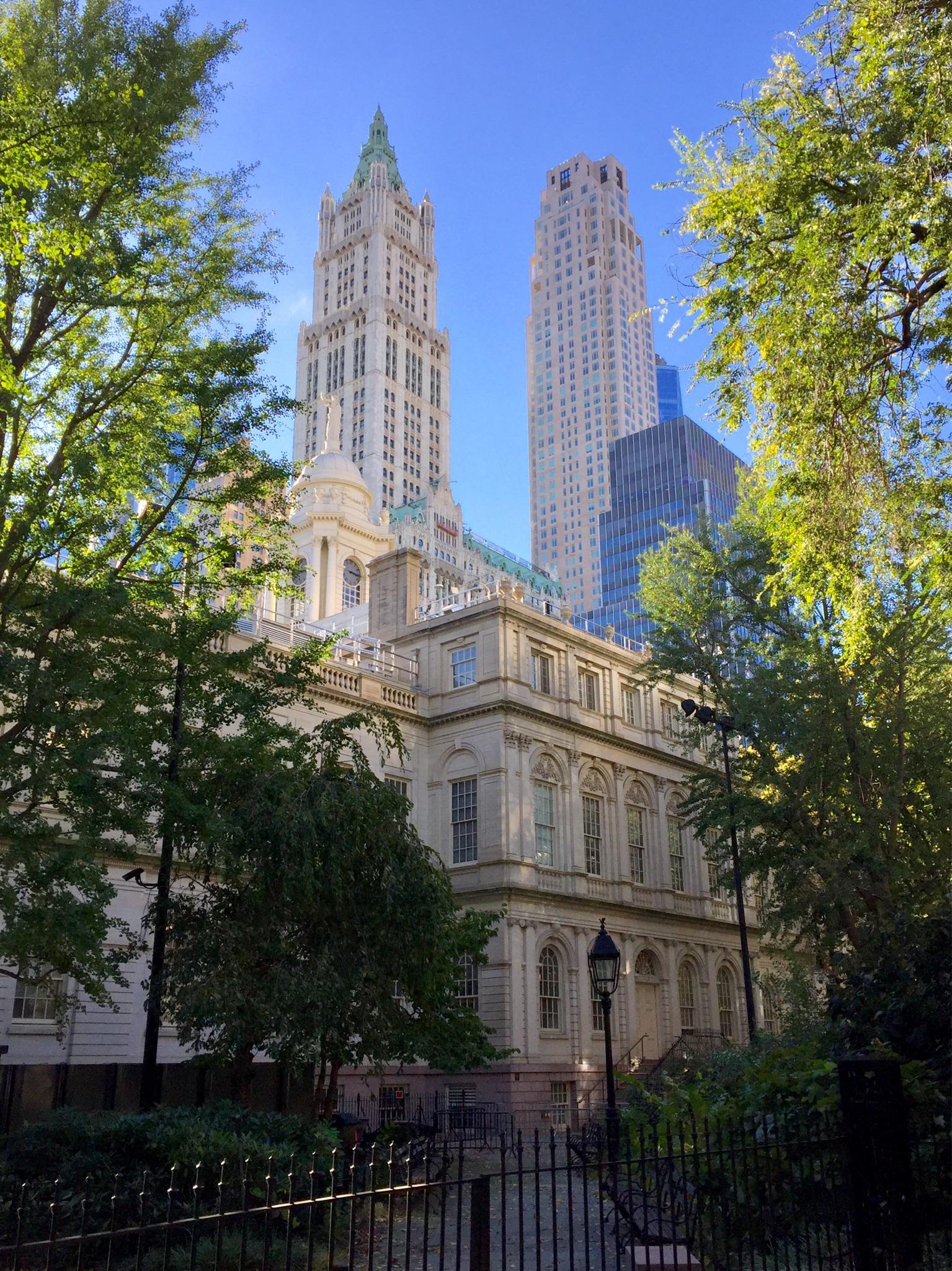 Historic building framed by trees, with modern skyscrapers behind under bright sun.