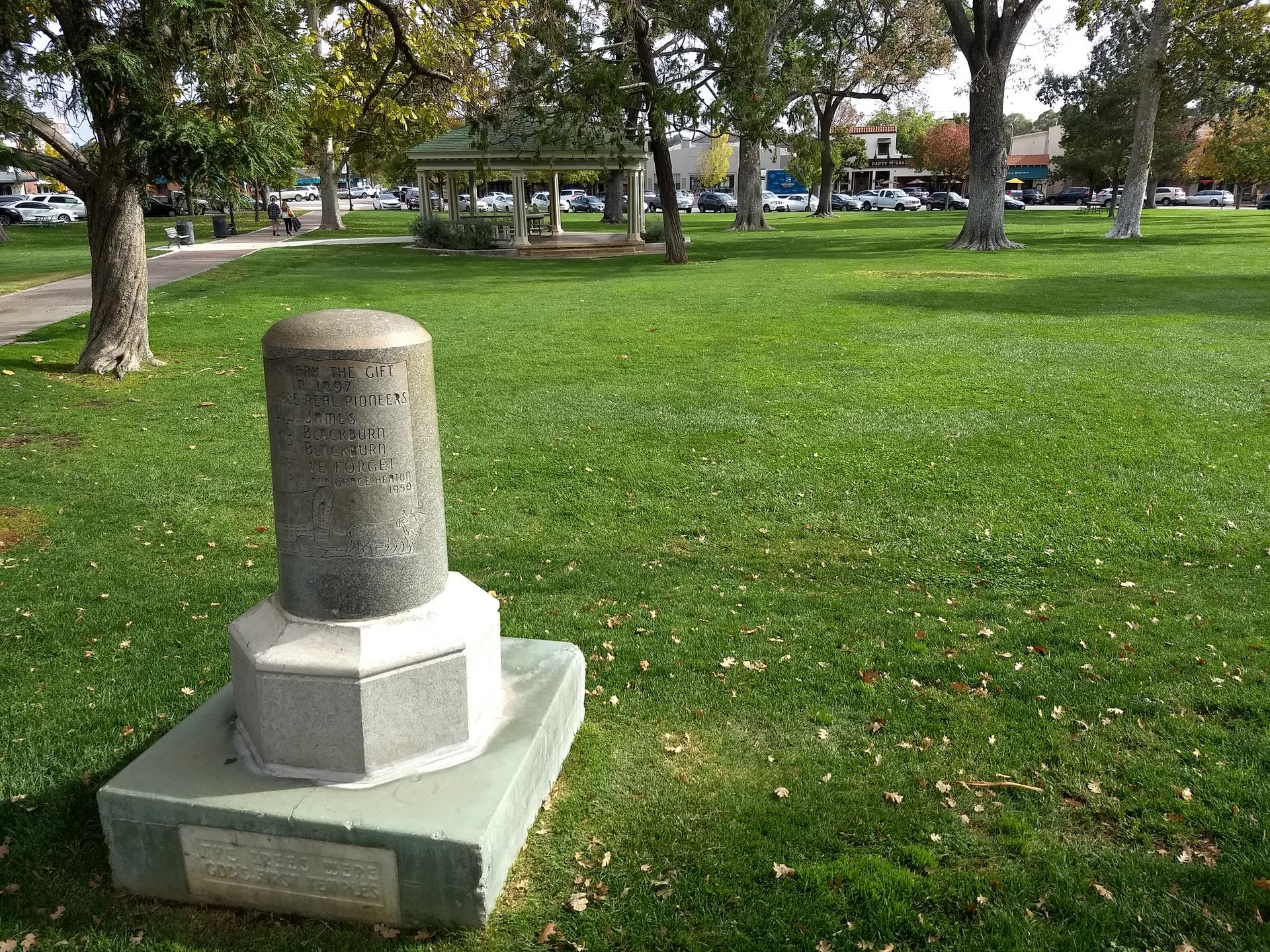 A stone monument stands on a green lawn in a sunny park with trees.