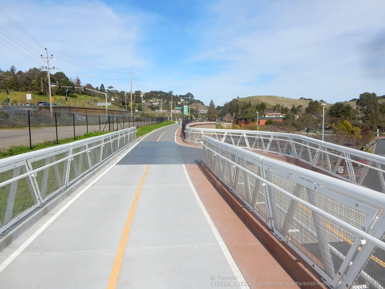 A paved path with white railings curves through a sunny, hilly landscape.