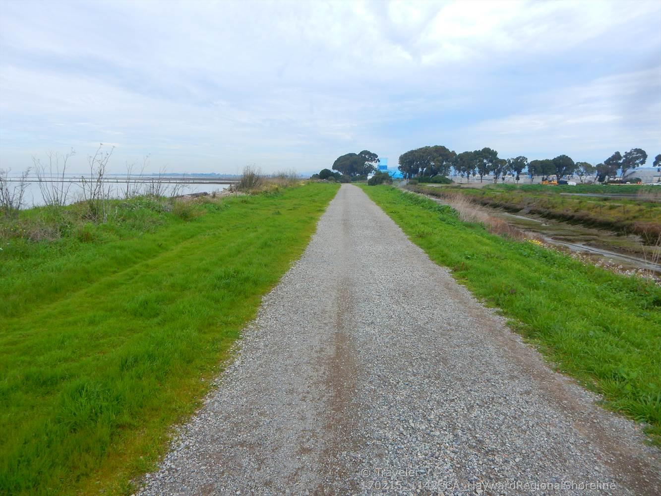 A gravel path extends through green grass towards distant trees under a cloudy sky.