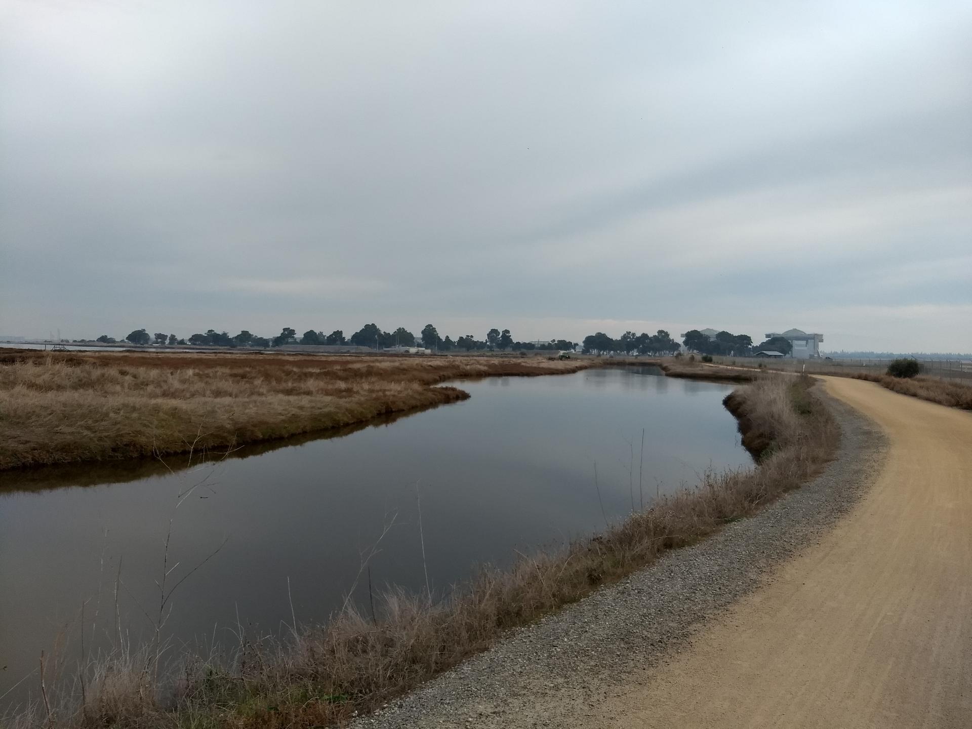 A calm waterway winds through dry fields next to a dirt path under a cloudy sky.
