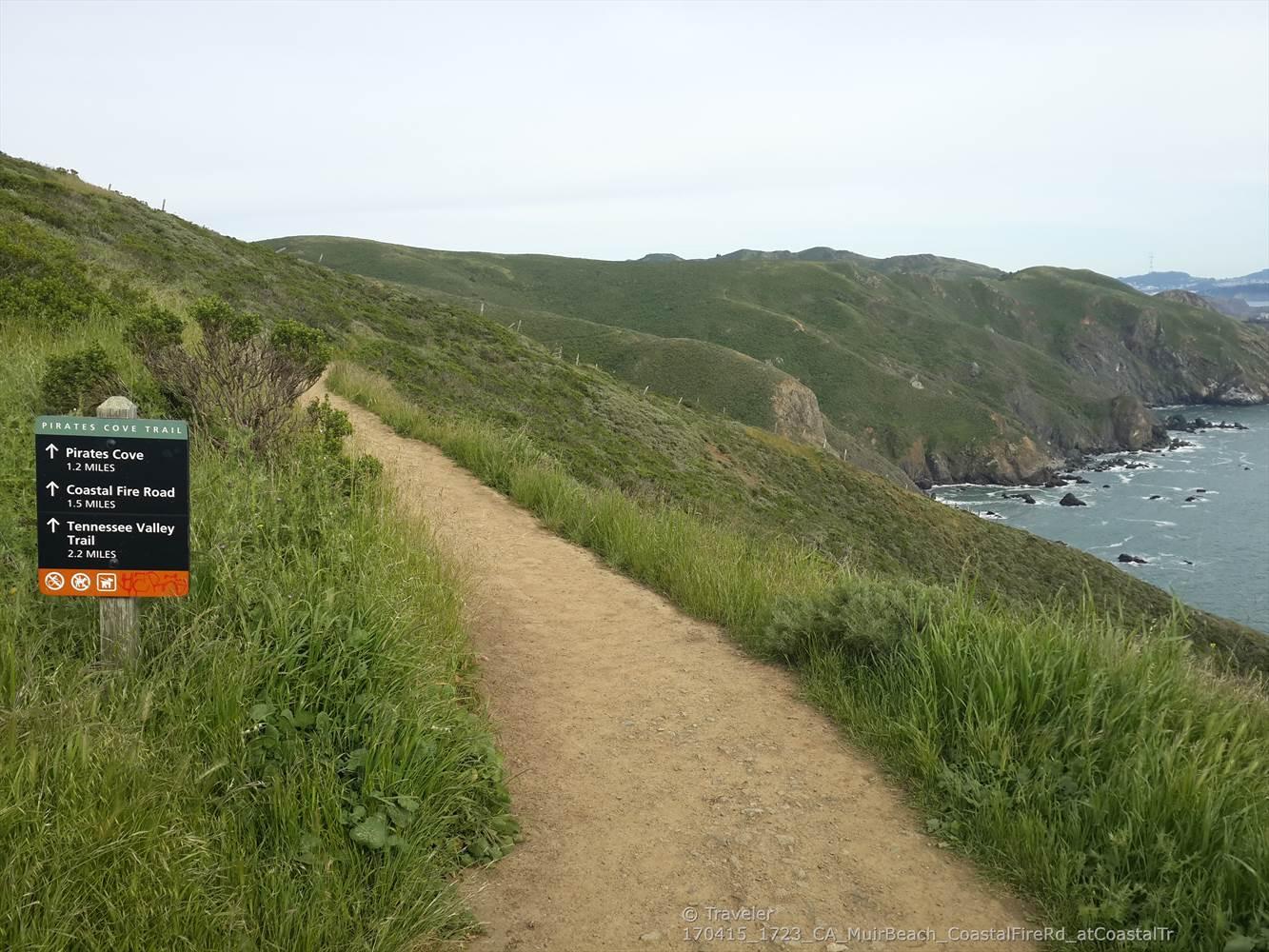 A trail sign stands beside a dirt path on a grassy coastal cliff.