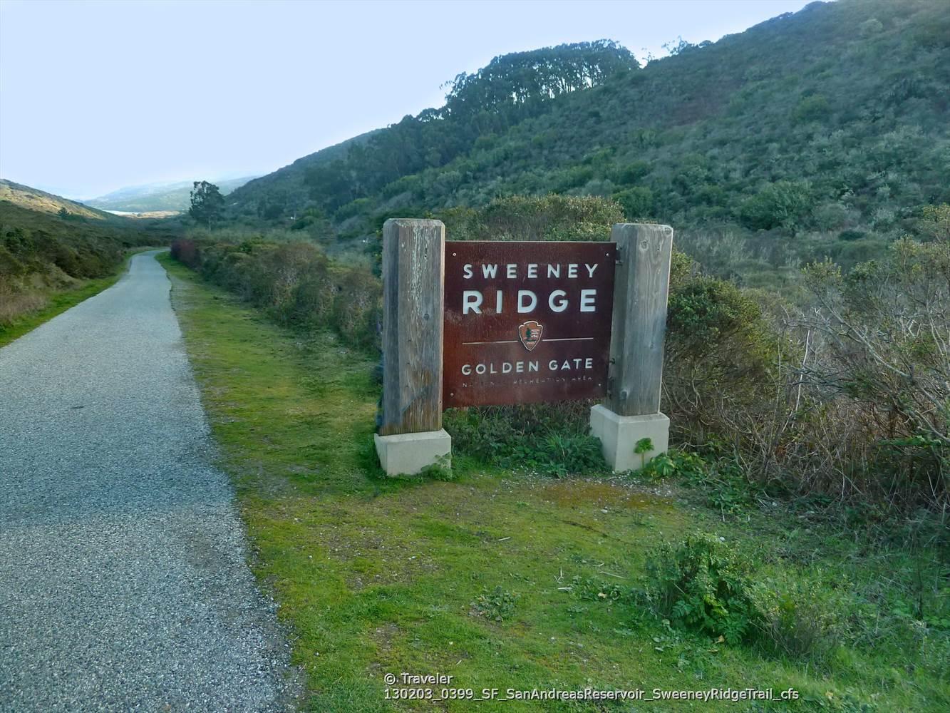 Wooden sign next to a paved path, surrounded by green hills.