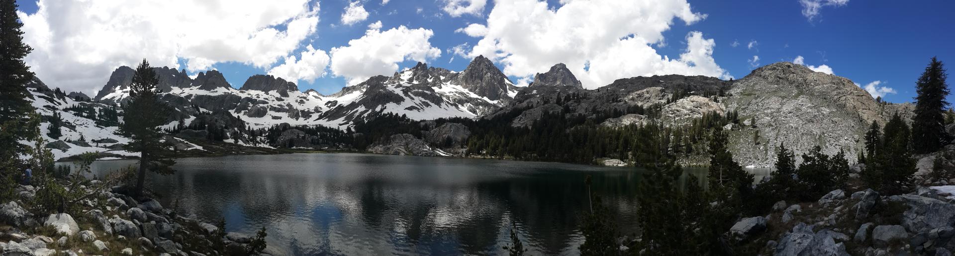 Panoramic view of a calm lake reflecting snow-capped jagged mountains under a cloudy sky.