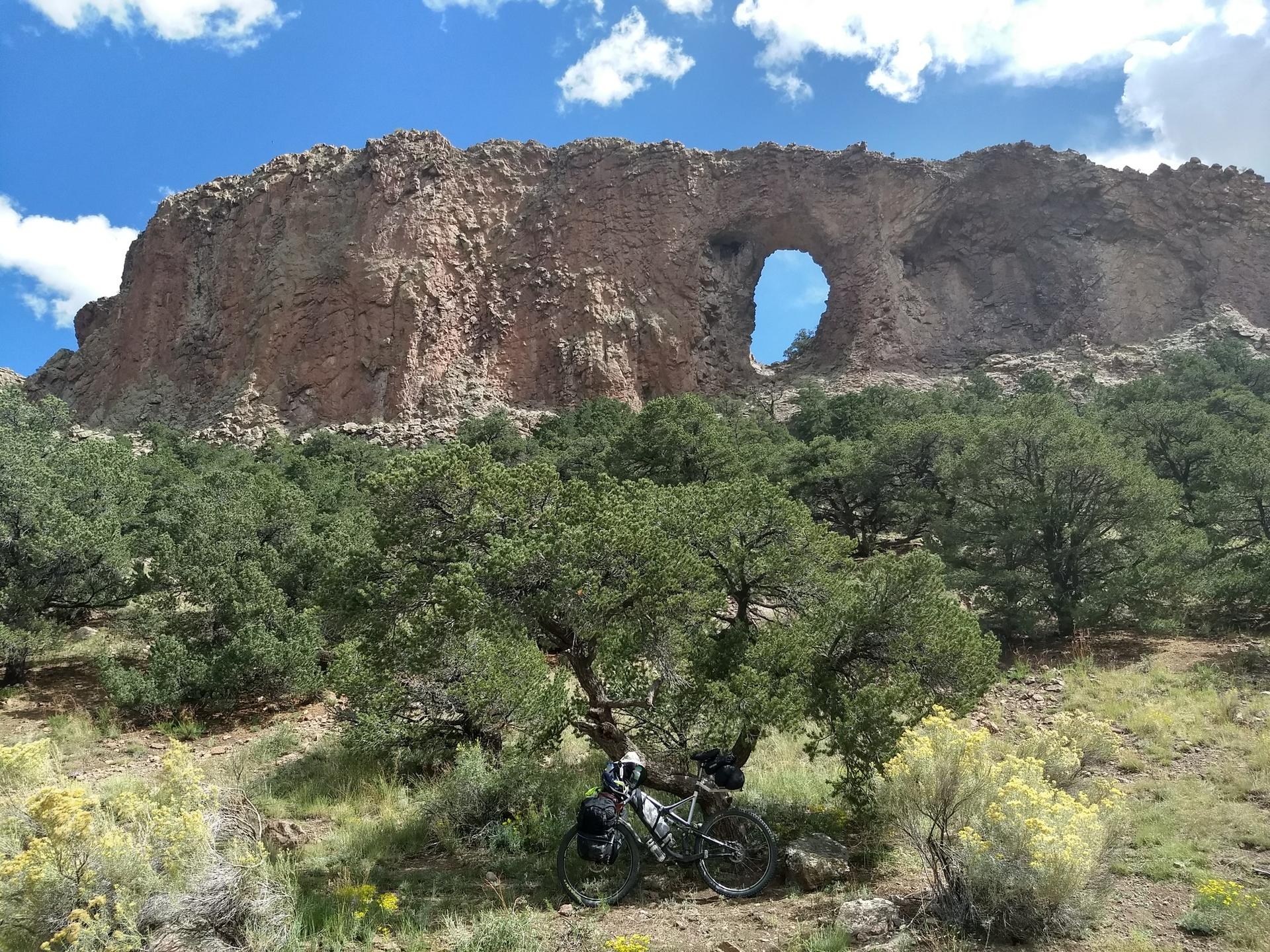 A bicycle rests under trees below a large rock arch on a sunny day.