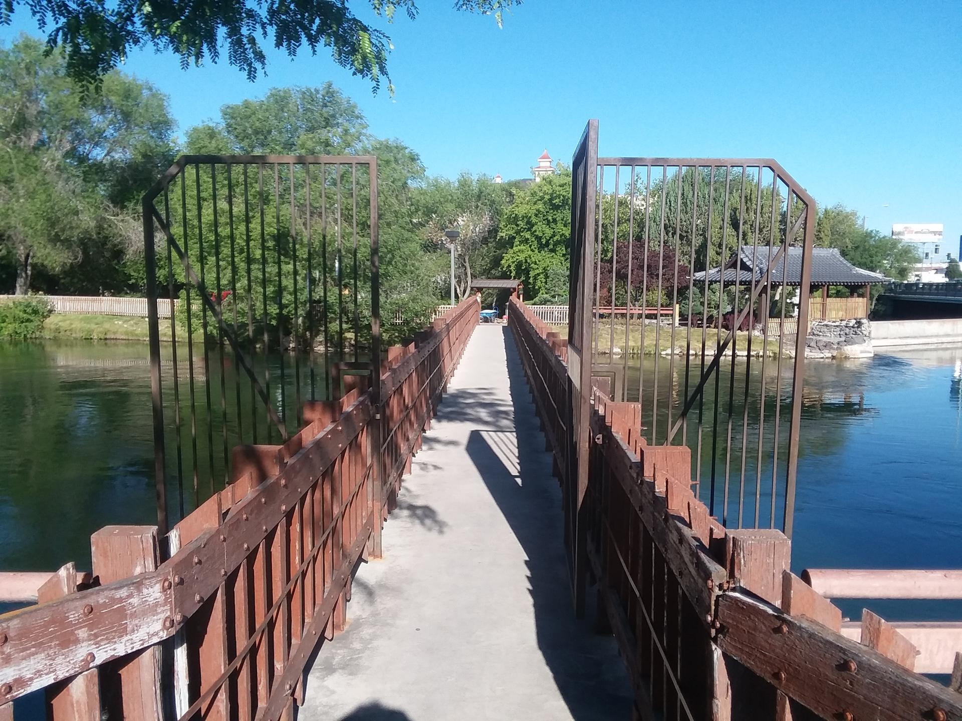 Wooden pedestrian bridge over a calm river, surrounded by green trees.