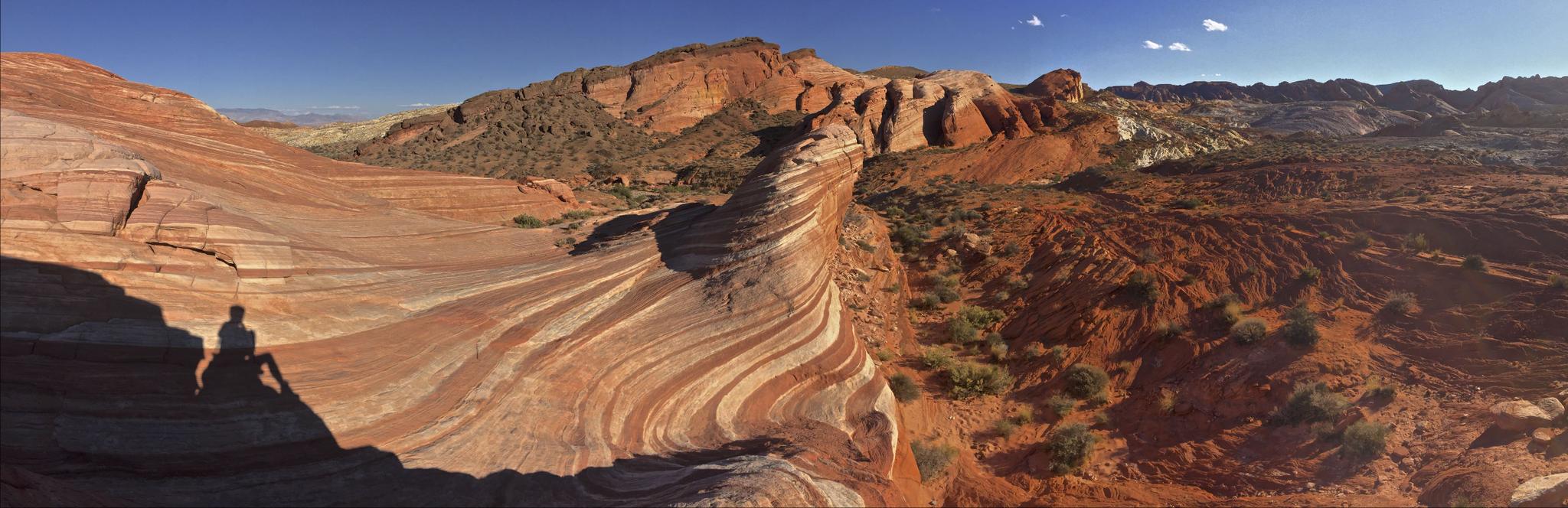 Person sitting on wavy red rock, panoramic desert landscape, bright sun.