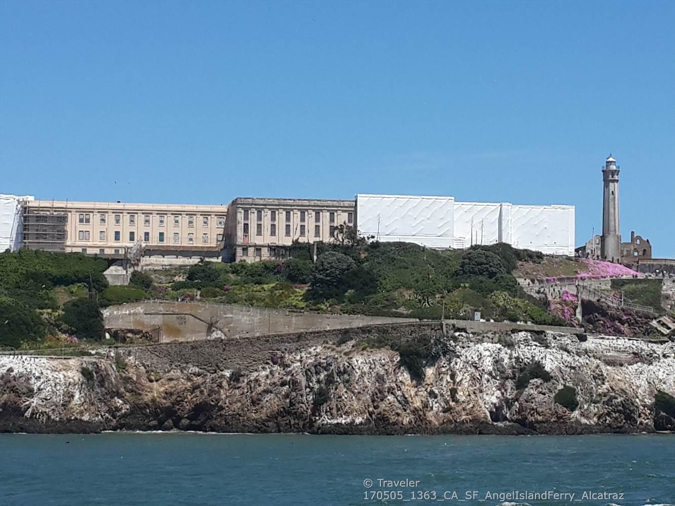 Large building complex and lighthouse on a rocky island, calm blue water.