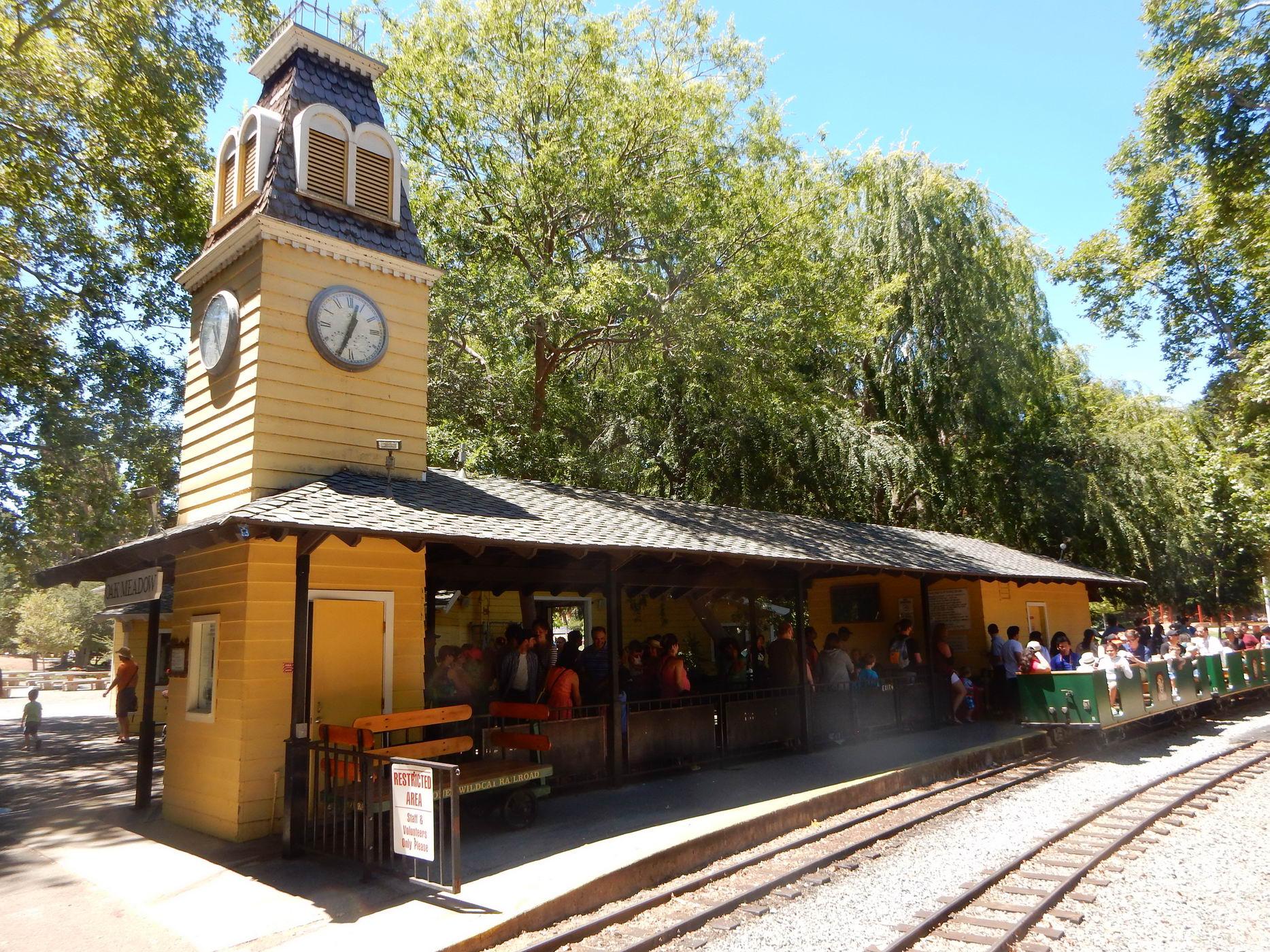 Yellow train station with a clock tower, people waiting for a train on a sunny day.
