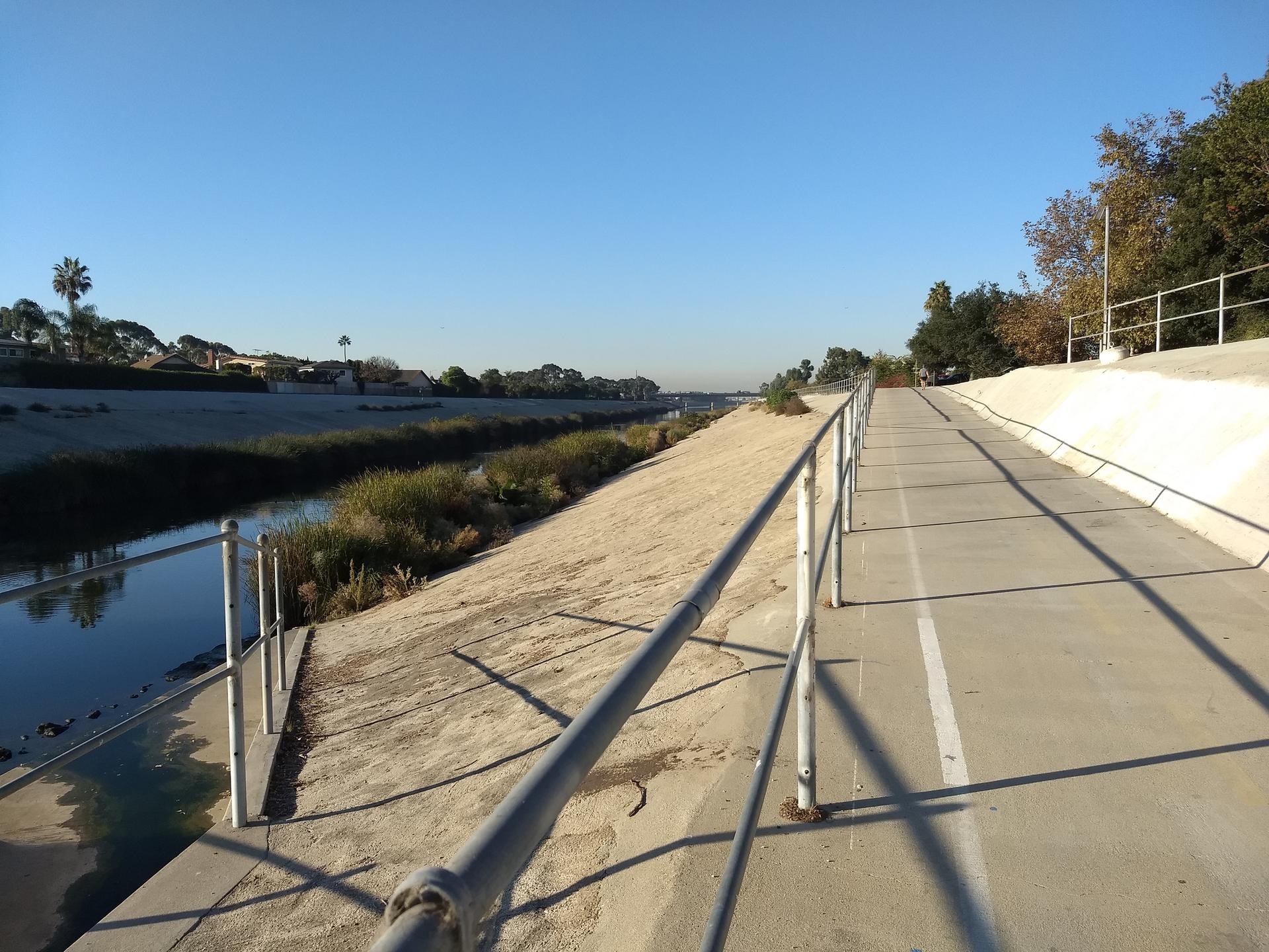 A concrete path with a metal railing alongside a canal under a clear sky.