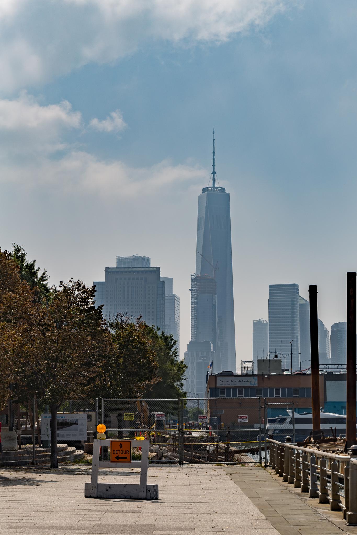 A very tall skyscraper dominates the hazy city skyline from a waterfront path.