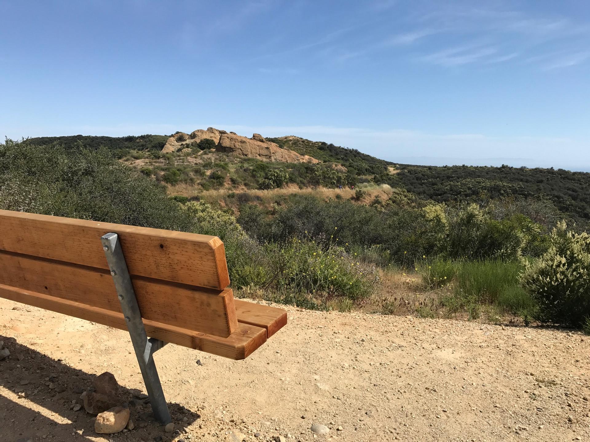 Wooden bench on a dirt path overlooking a sunny, brush-covered landscape.