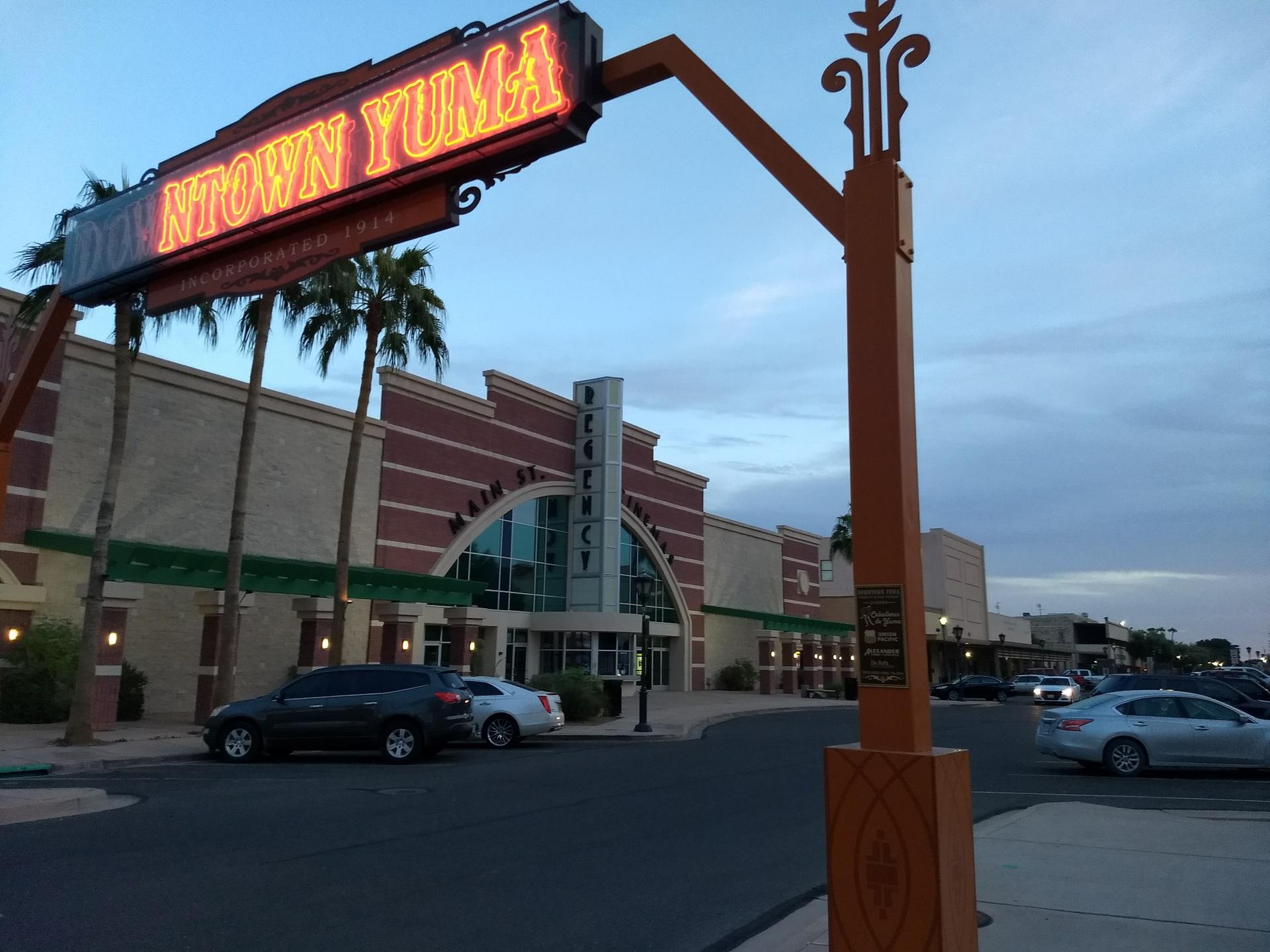 Neon sign over a street with cars and buildings at dusk.