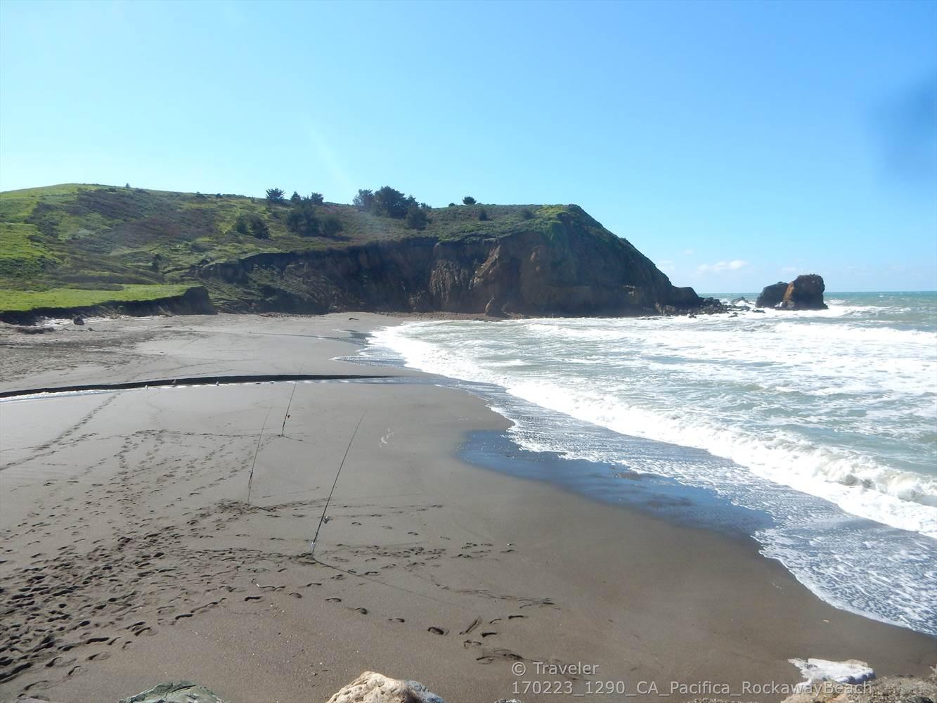 Waves crash on a sandy beach next to steep, green-topped cliffs.