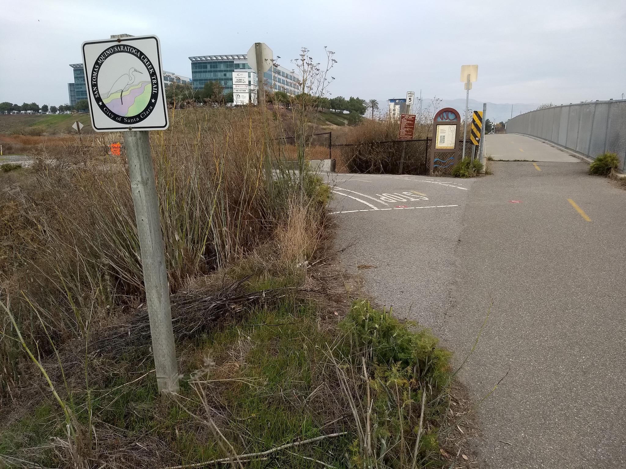 A trail sign stands by a paved path and dry brush under an overcast sky.