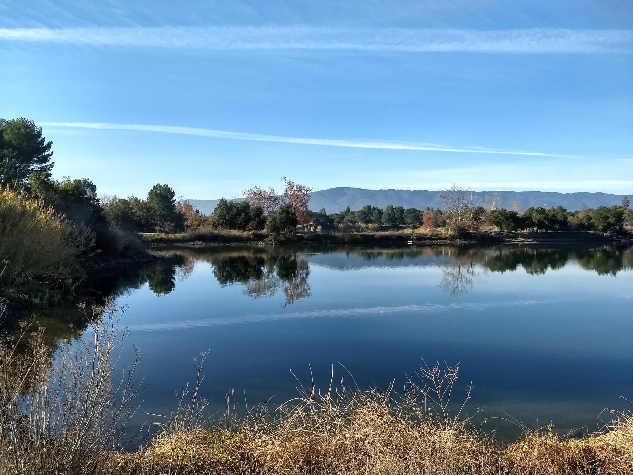 Calm lake reflecting trees and mountains under a clear blue sky.