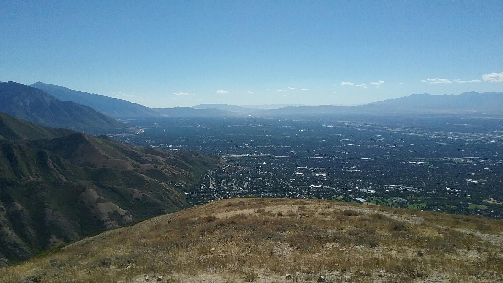 Wide view of a sprawling city and hazy mountains from a dry, grassy hill.