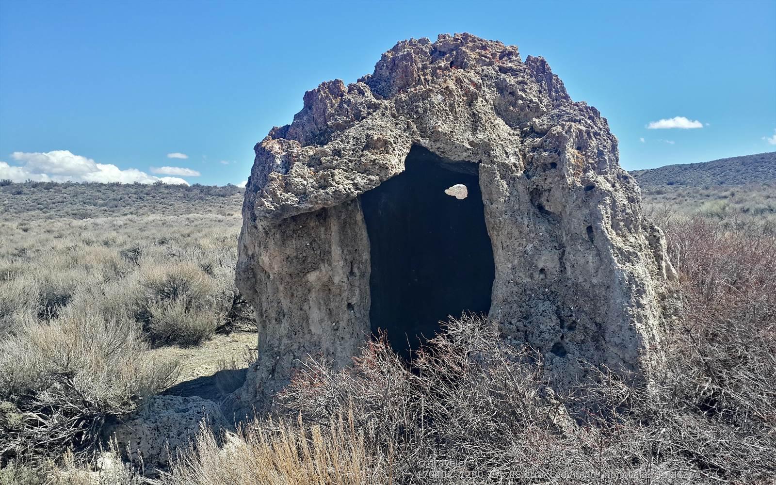 A large, hollow rock formation stands in an arid, brushy landscape under a clear sky.