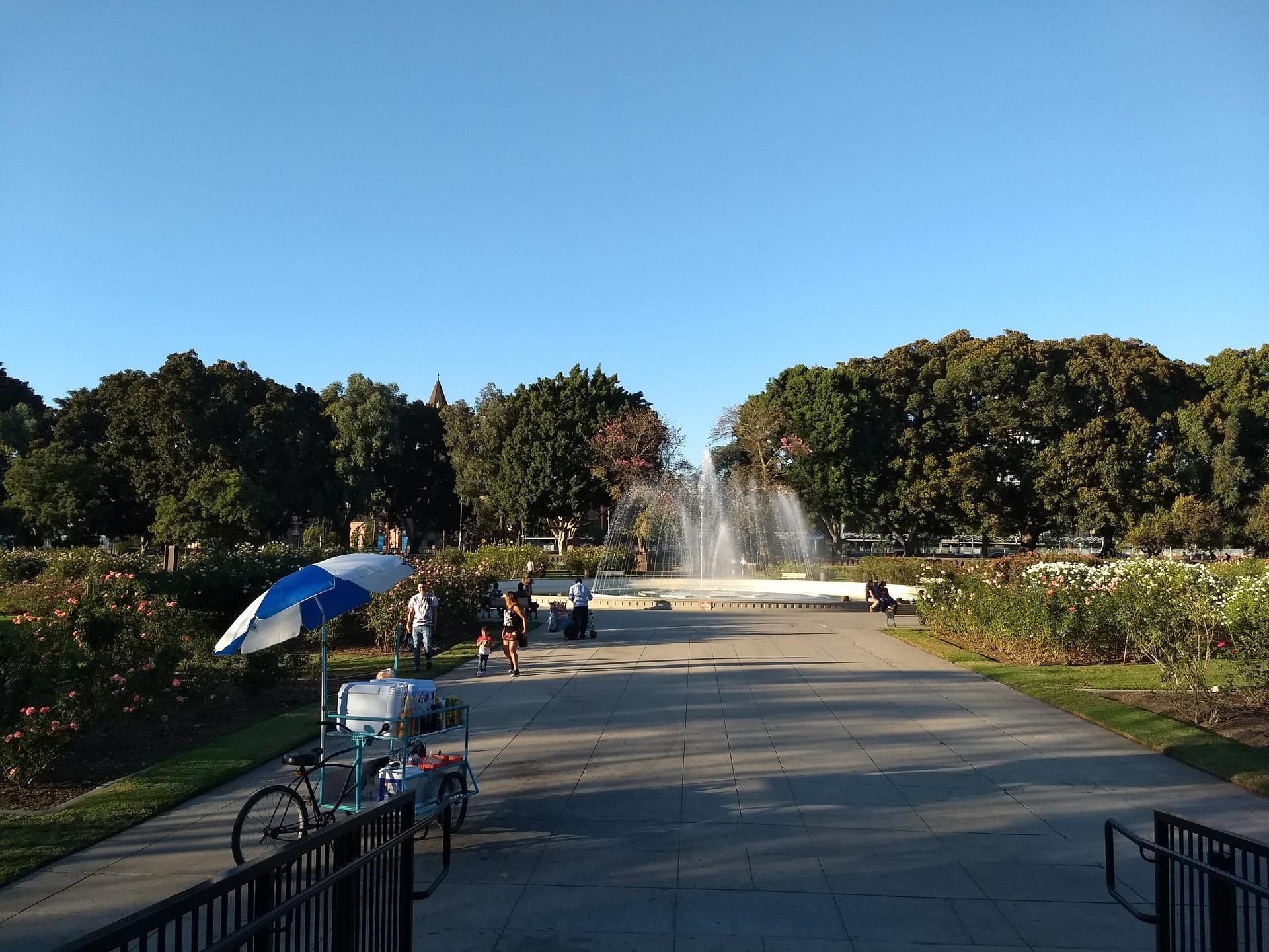 Sunny park with a fountain, vendor cart, and people walking.