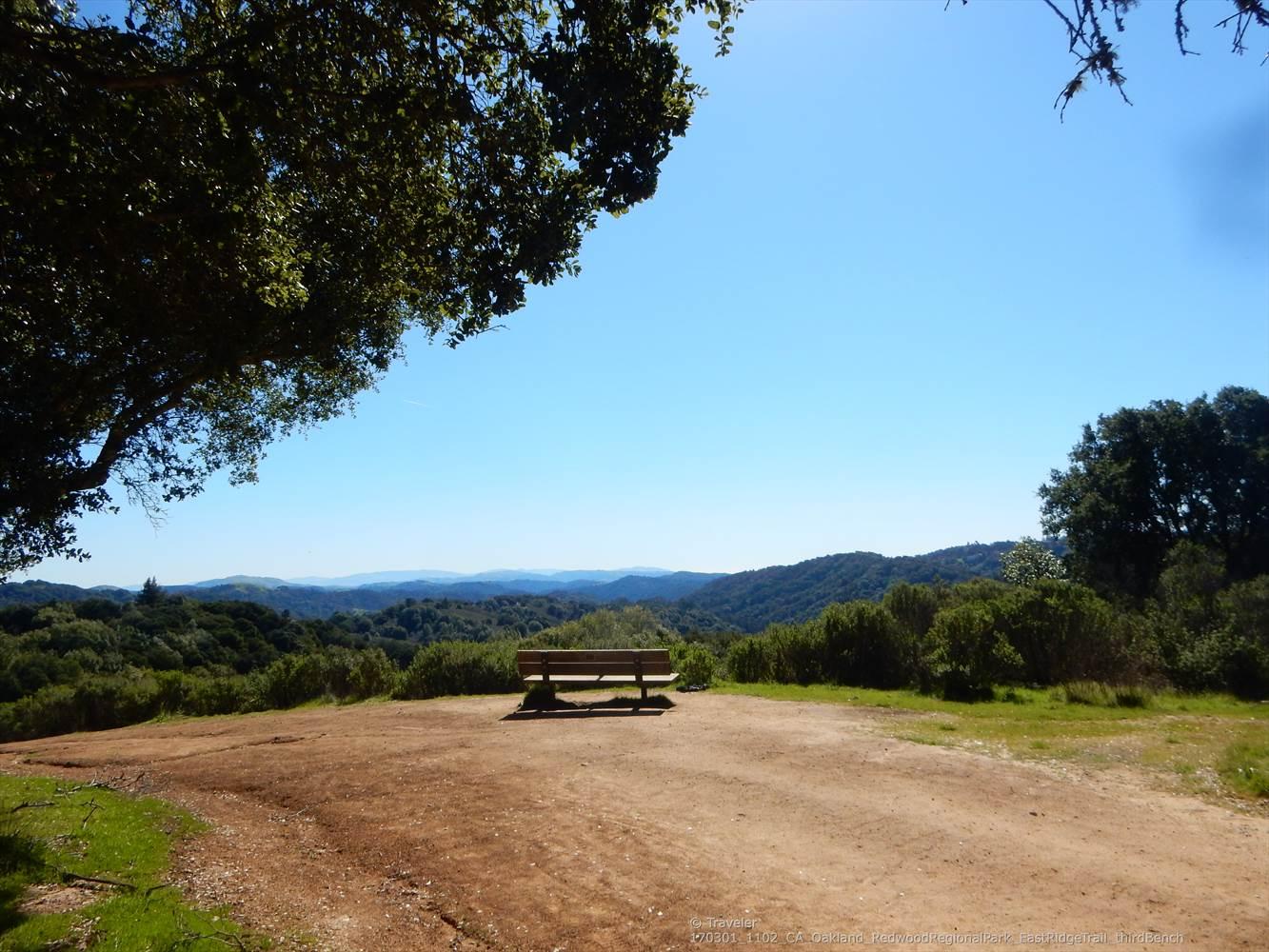 Wooden bench on a dirt clearing overlooking a sunny, forested valley.