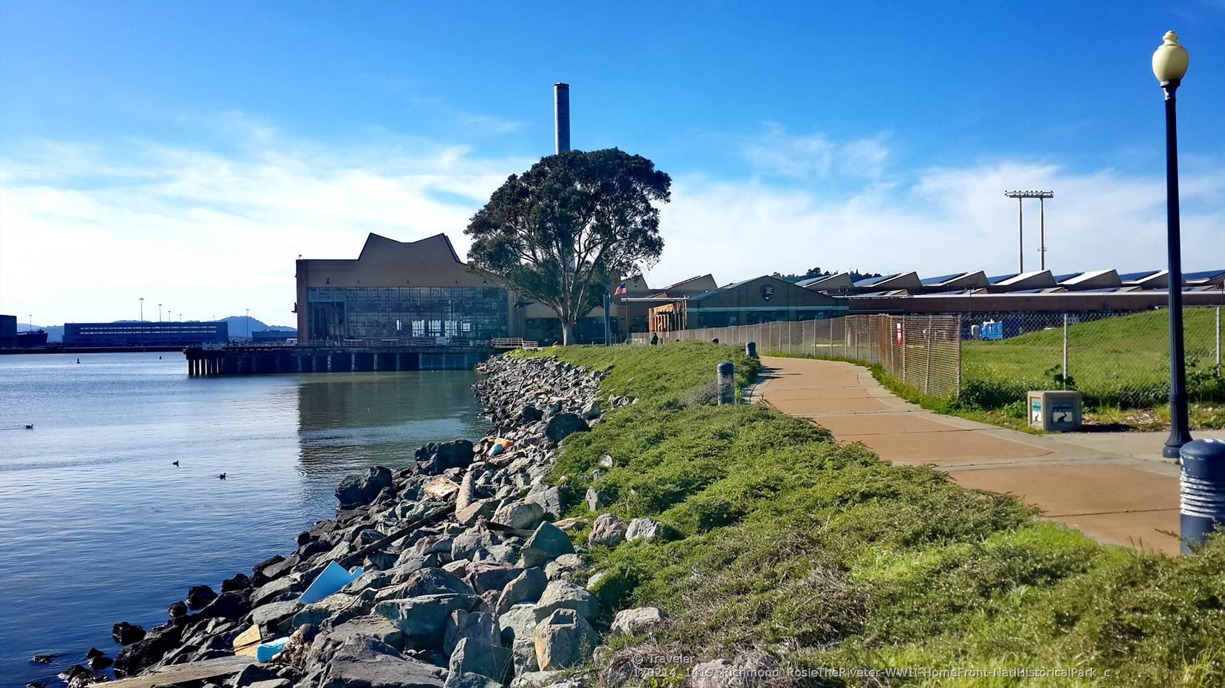 Paved path along a rocky waterfront with industrial buildings and a tall chimney.