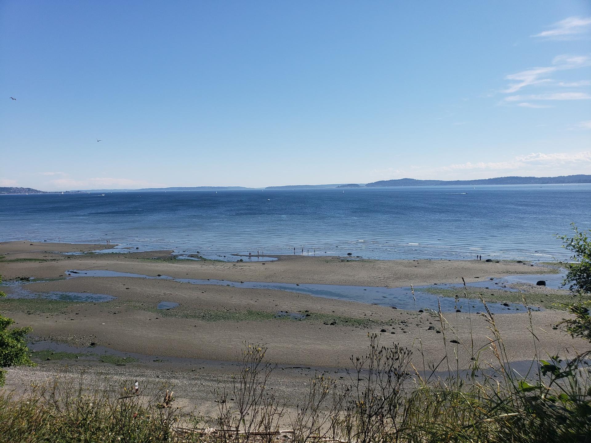 Wide sandy beach at low tide, calm blue water, and distant land.