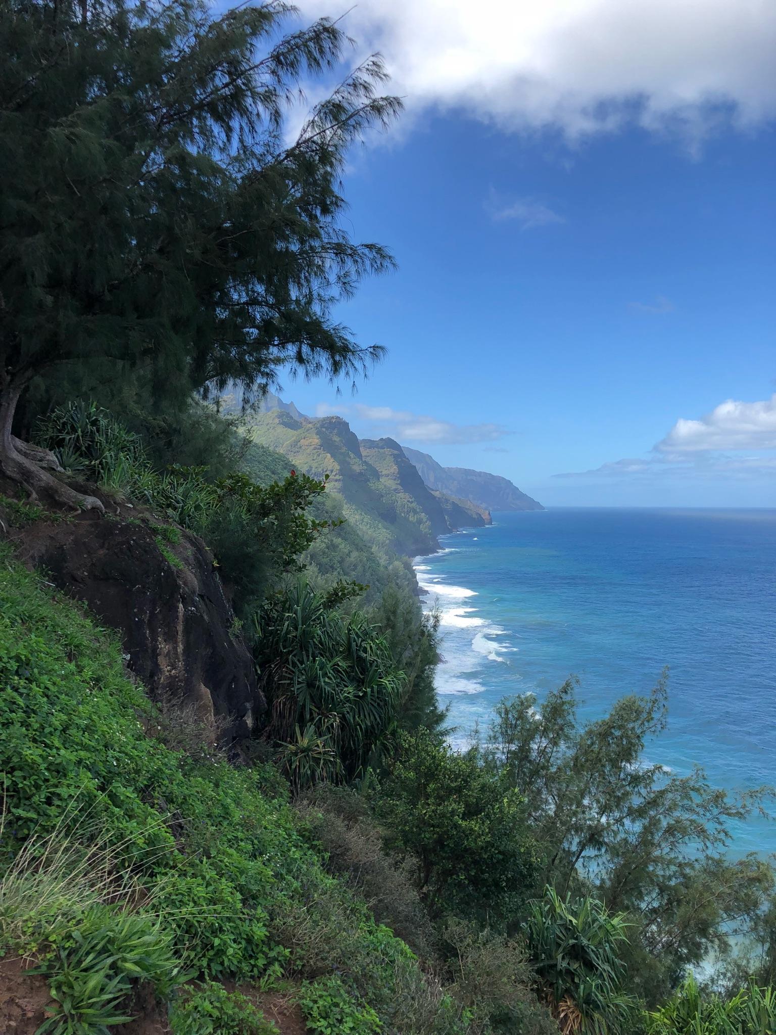 Steep green mountainside overlooking a bright blue ocean with white waves.
