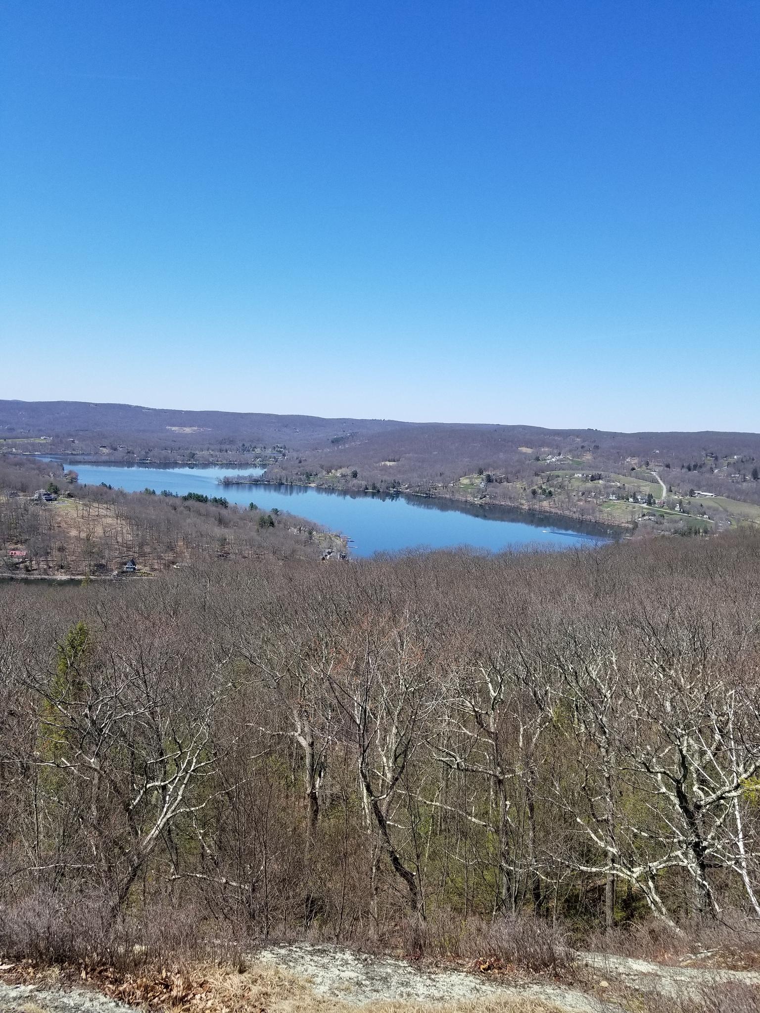 Expansive view of a lake surrounded by bare trees and distant hills under clear sky.