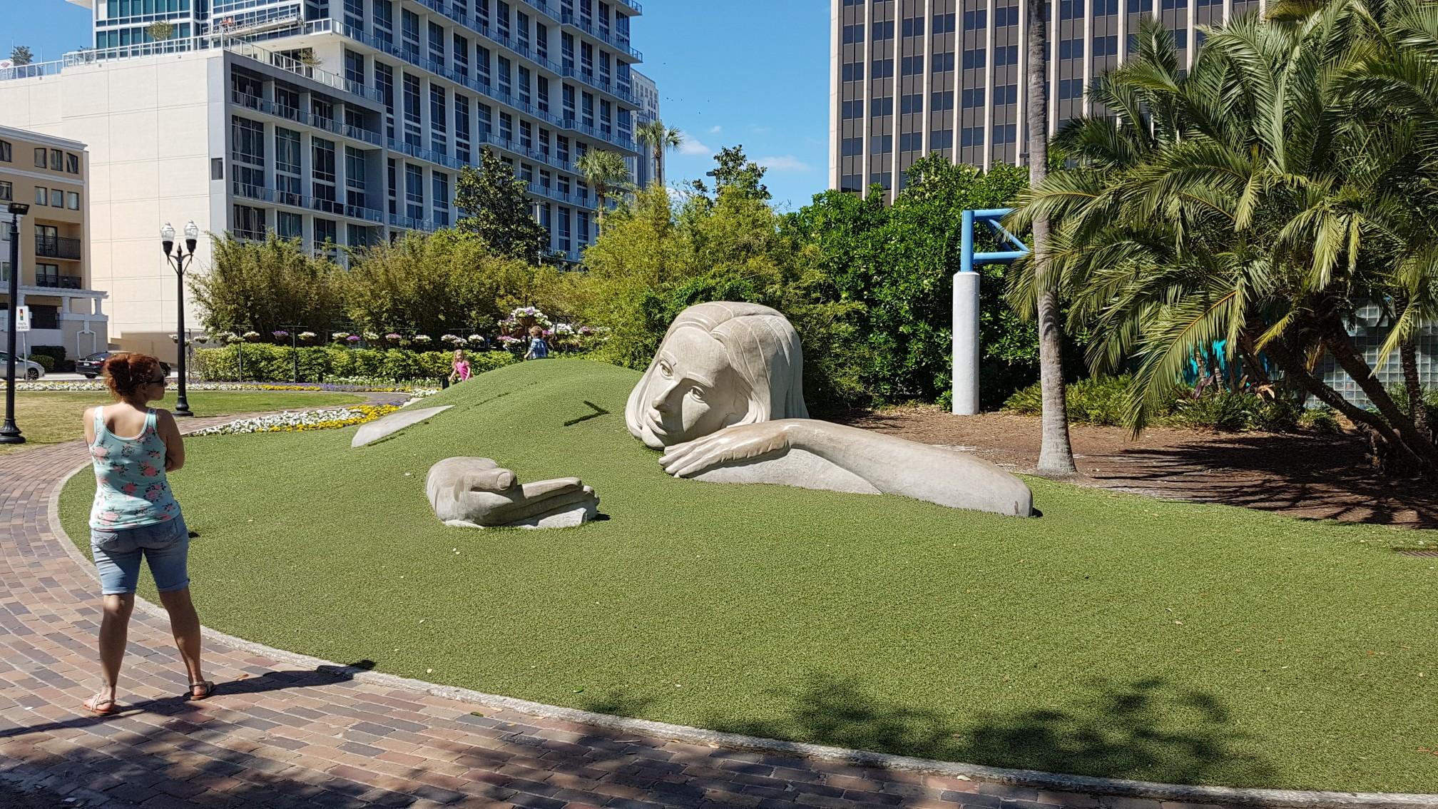 Woman views large sculpture of a reclining figure in a sunny park.