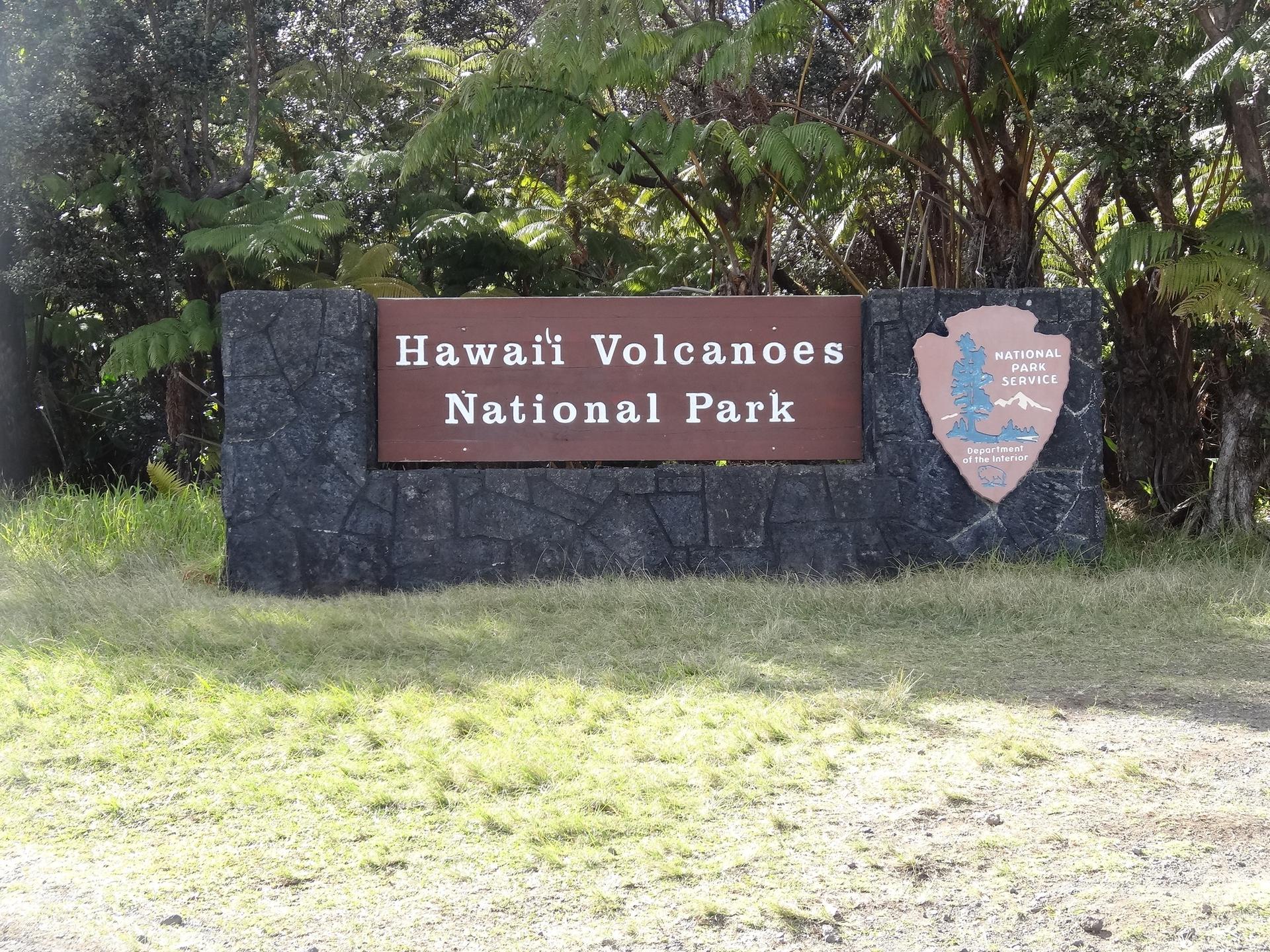 A brown park sign on a stone base, with green trees behind it.