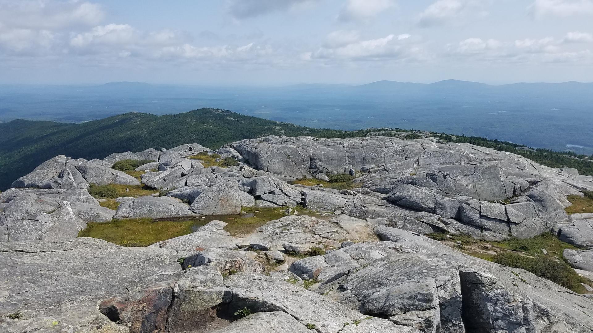 Rocky mountain summit with distant green hills under a partly cloudy sky.