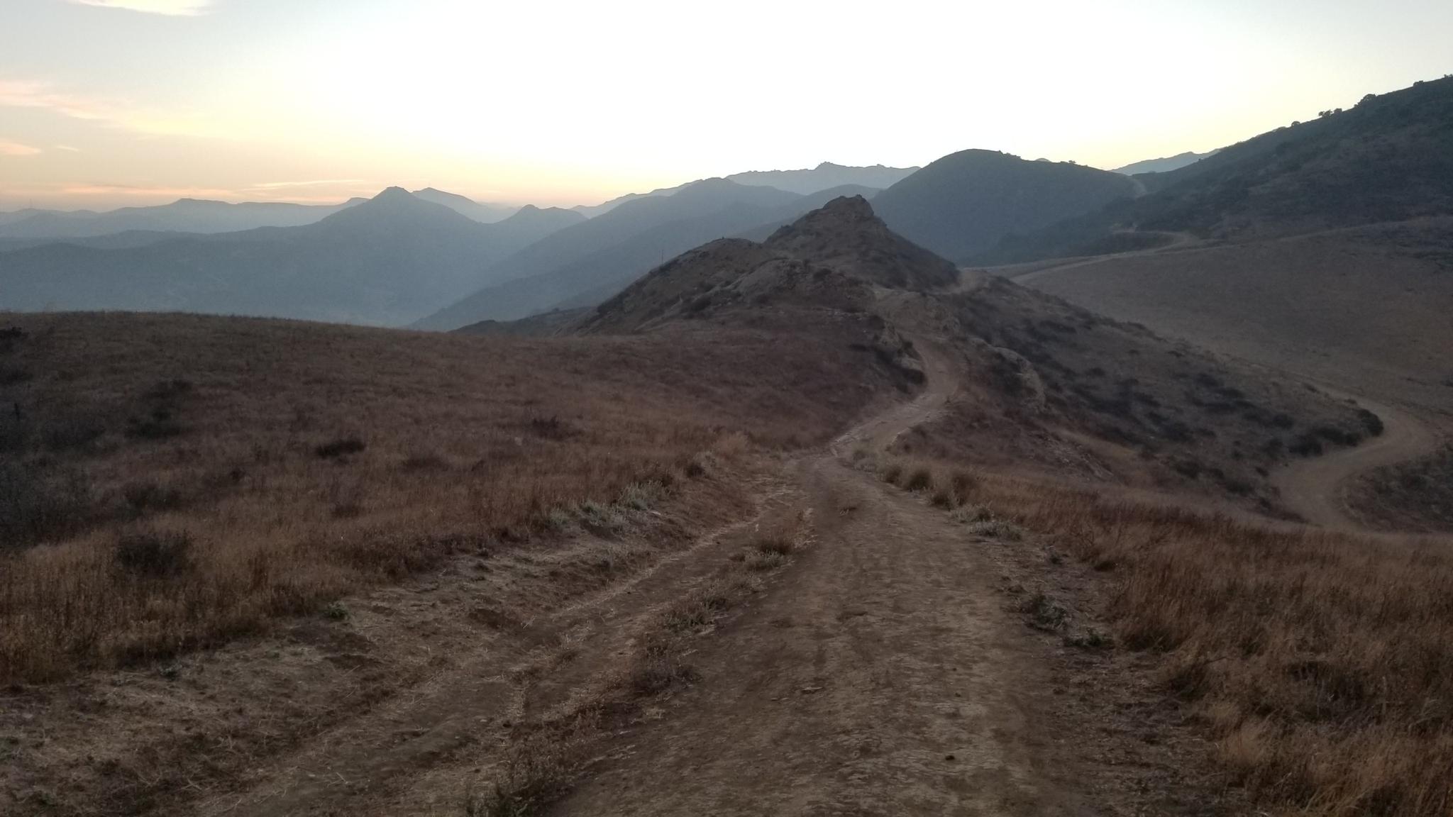 Dirt path winding through dry hills with distant mountains at dawn.