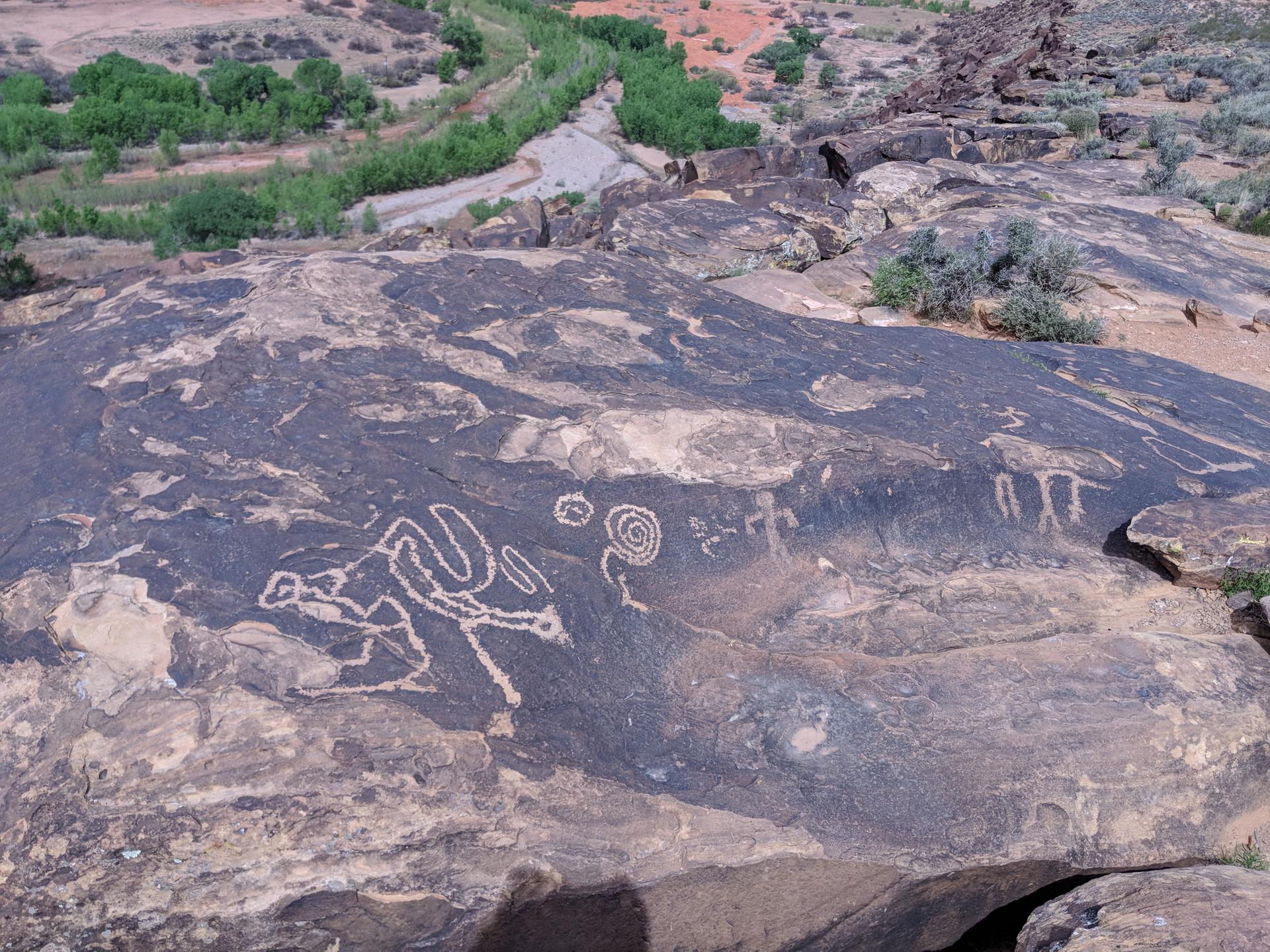 Dark rock with petroglyphs, overlooking a green desert valley in daylight.
