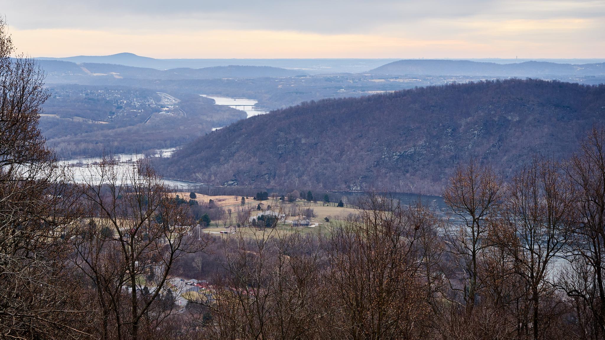 Overcast view of a wide river valley with forested hills and distant mountains.