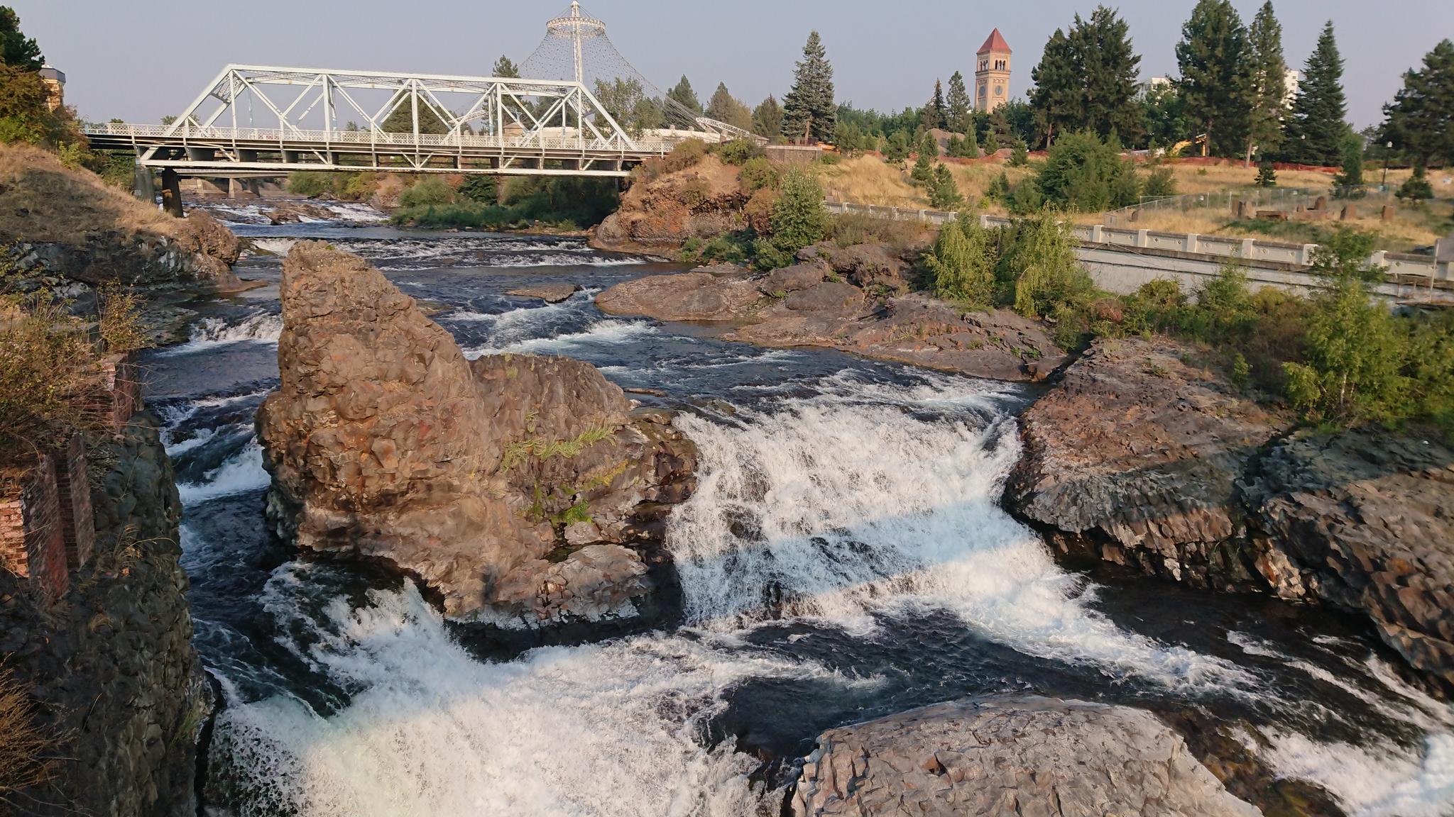 Sunny view of river rapids, a white truss bridge, and distant buildings.